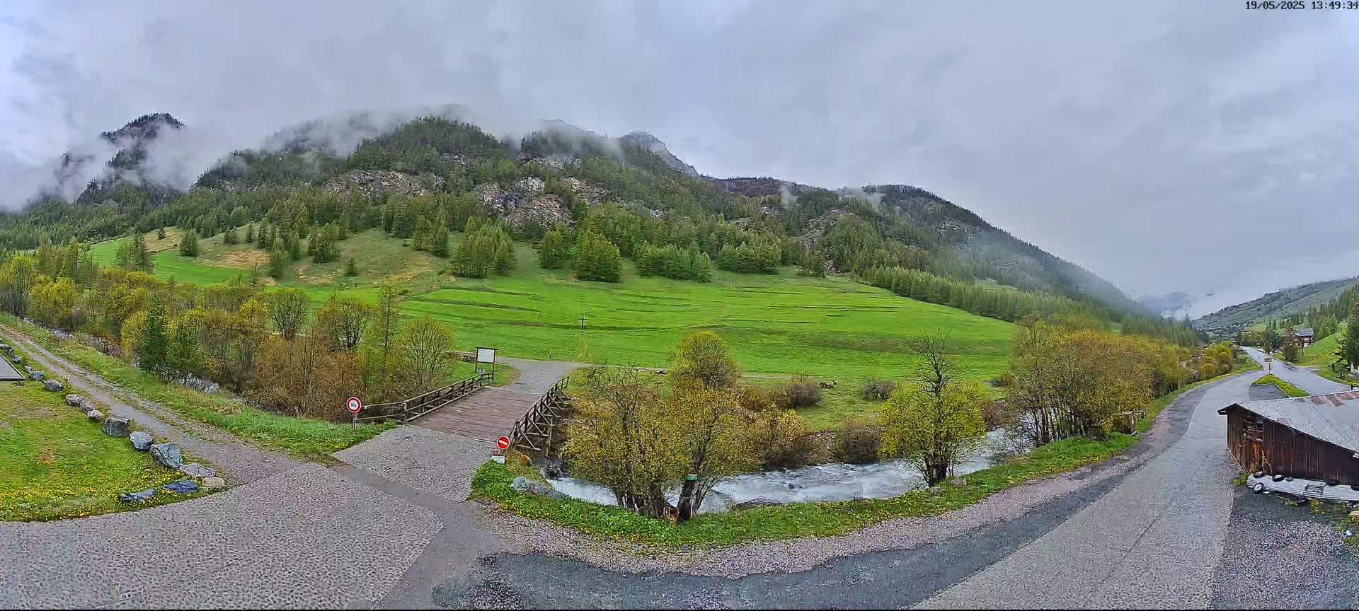 A small wooden bridge crosses a stream in a valley, with green fields and a mountain range shrouded in mist under an overcast sky.