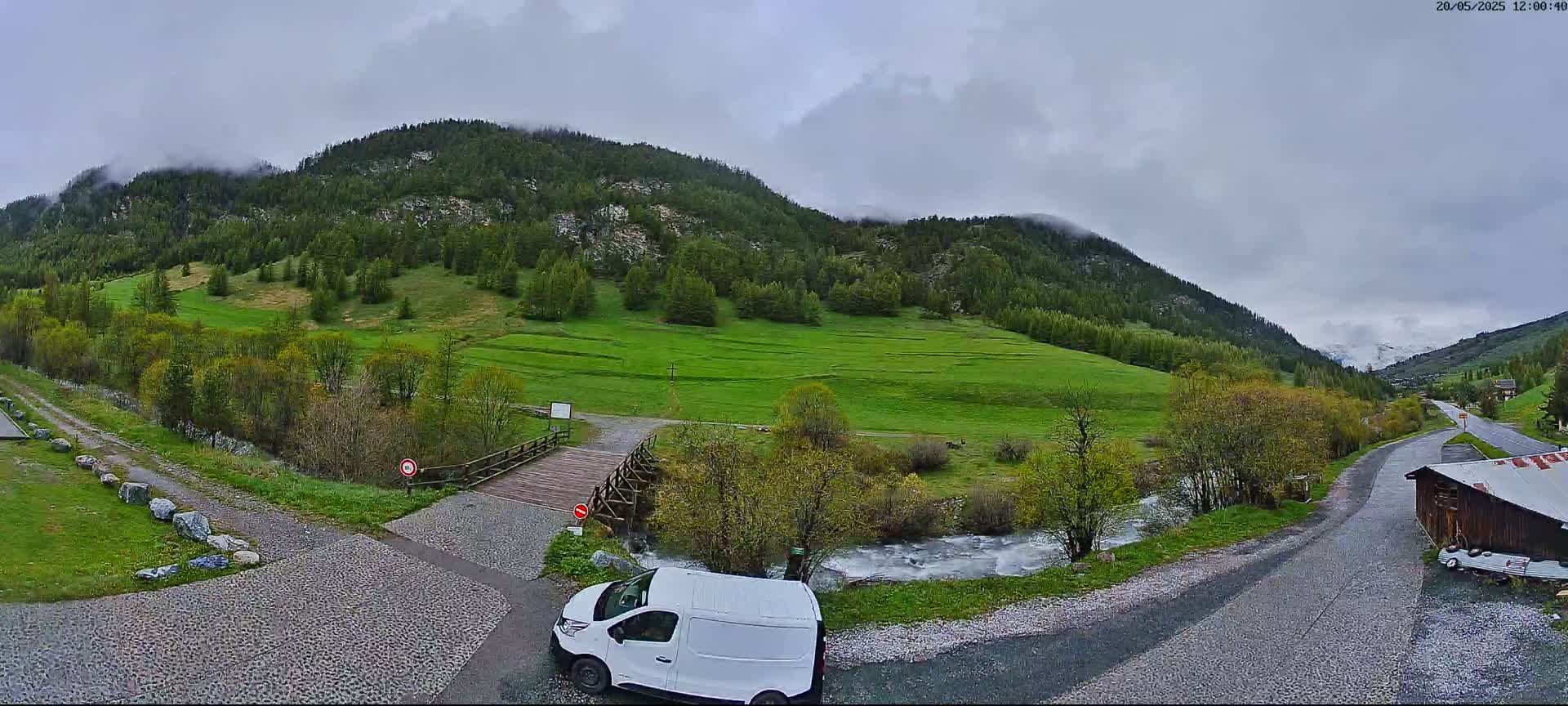 A white van is parked near a small wooden bridge over a stream in a green valley, surrounded by mountains under a cloudy sky.