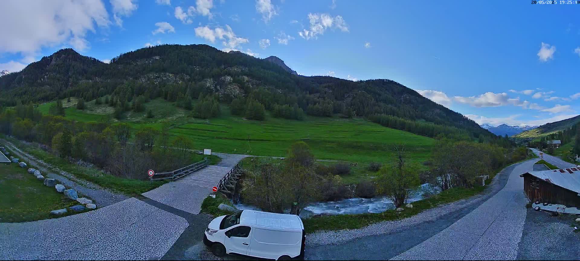 A white van is parked near a small wooden bridge over a stream, with green fields and mountains in the background under a mostly sunny sky.