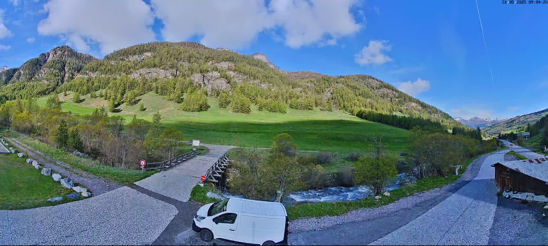 A white van is parked near a small wooden bridge over a stream, with a grassy valley and forested mountains in the background under a partly cloudy sky.