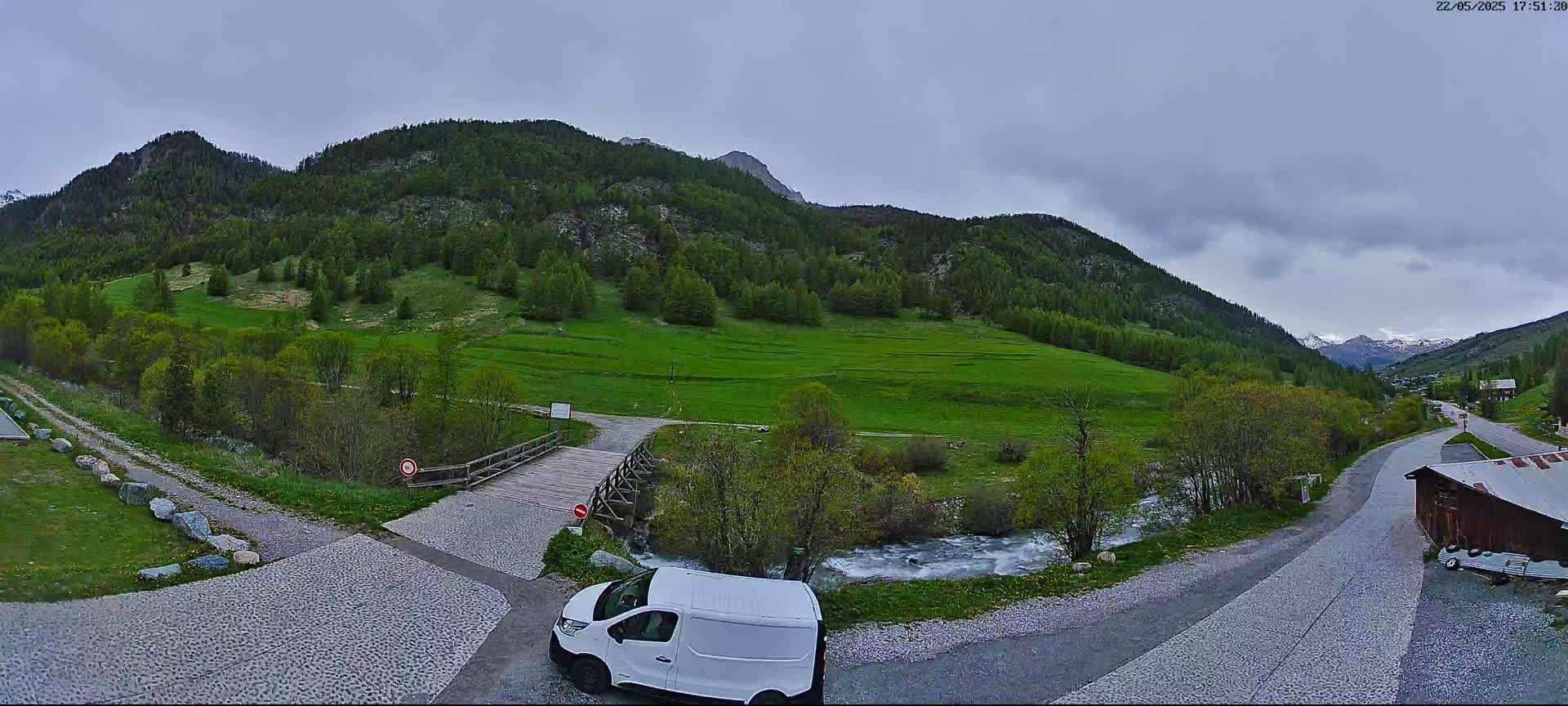 A white van is parked near a small wooden bridge over a stream, with green hills and mountains in the background under an overcast sky.