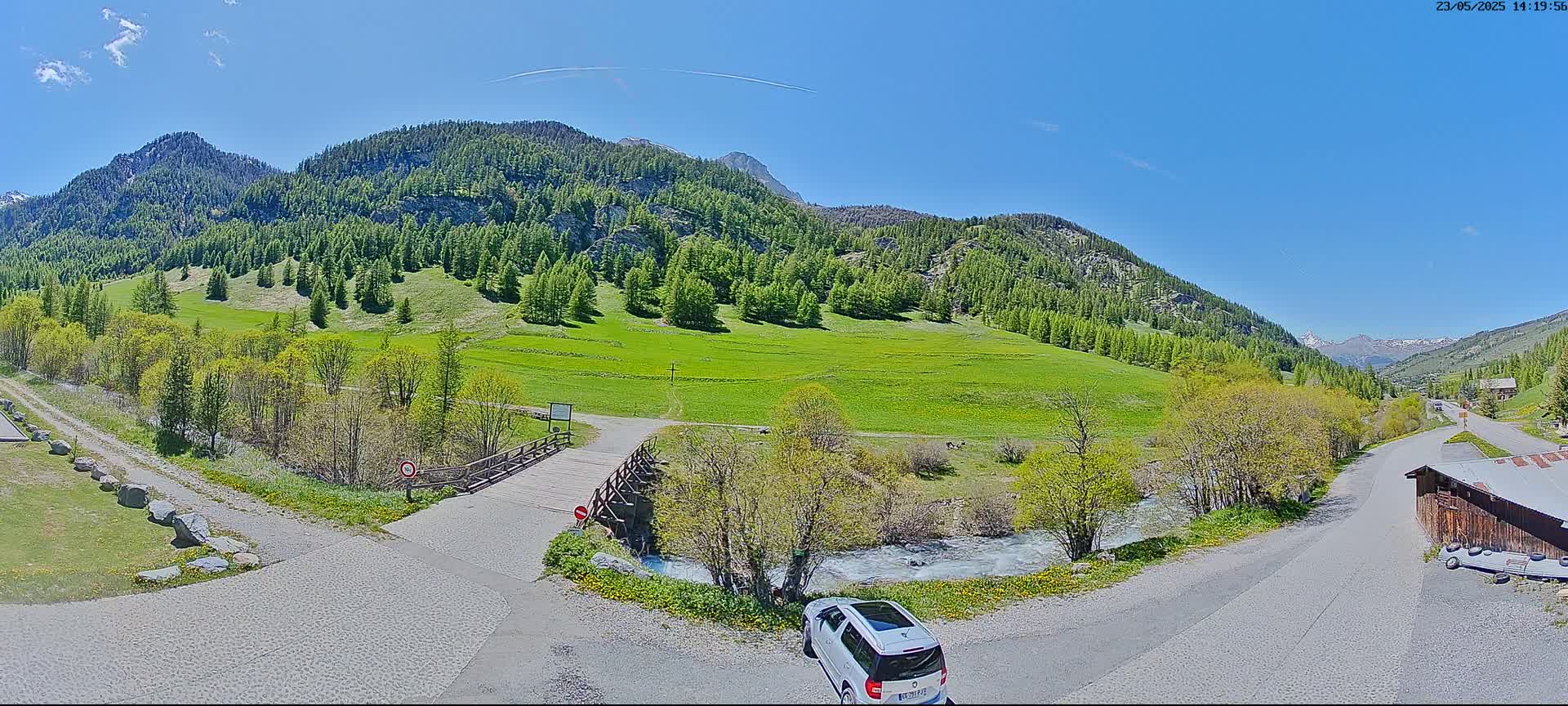 A white SUV is parked near a wooden bridge over a small stream in a sunny valley surrounded by green meadows and coniferous-covered mountains.