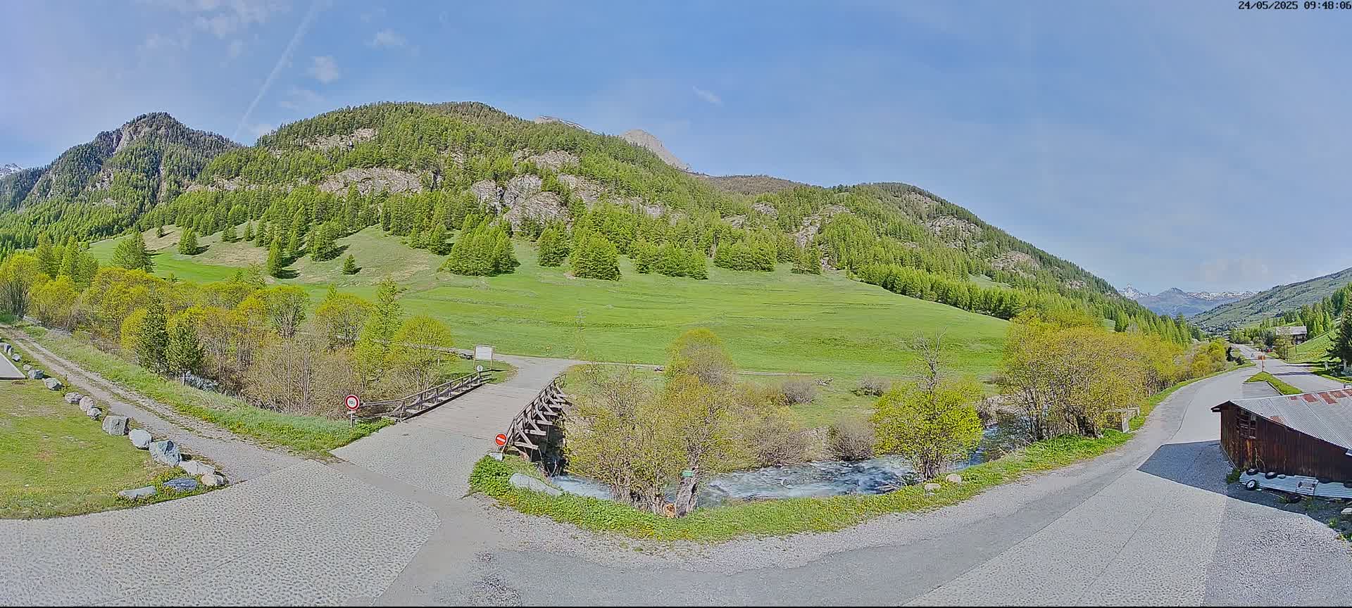 A sunny day reveals a mountain valley scene with a small wooden bridge crossing a stream, roads leading to a small building, and green meadows surrounded by evergreen trees on the mountain slopes.