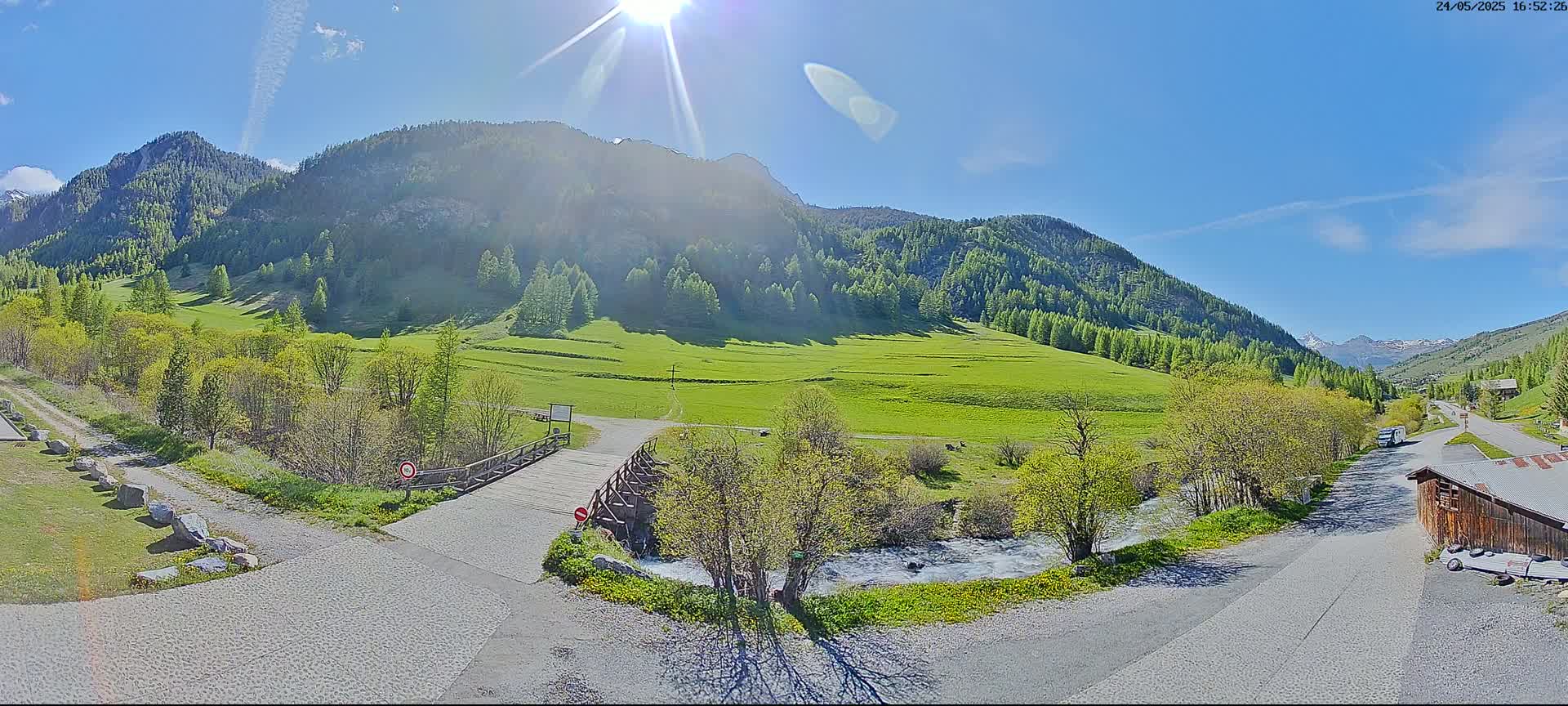 A sunny day in a mountainous valley reveals a small wooden bridge over a stream, leading to a grassy field and a road winding through green hillsides under a clear blue sky.
