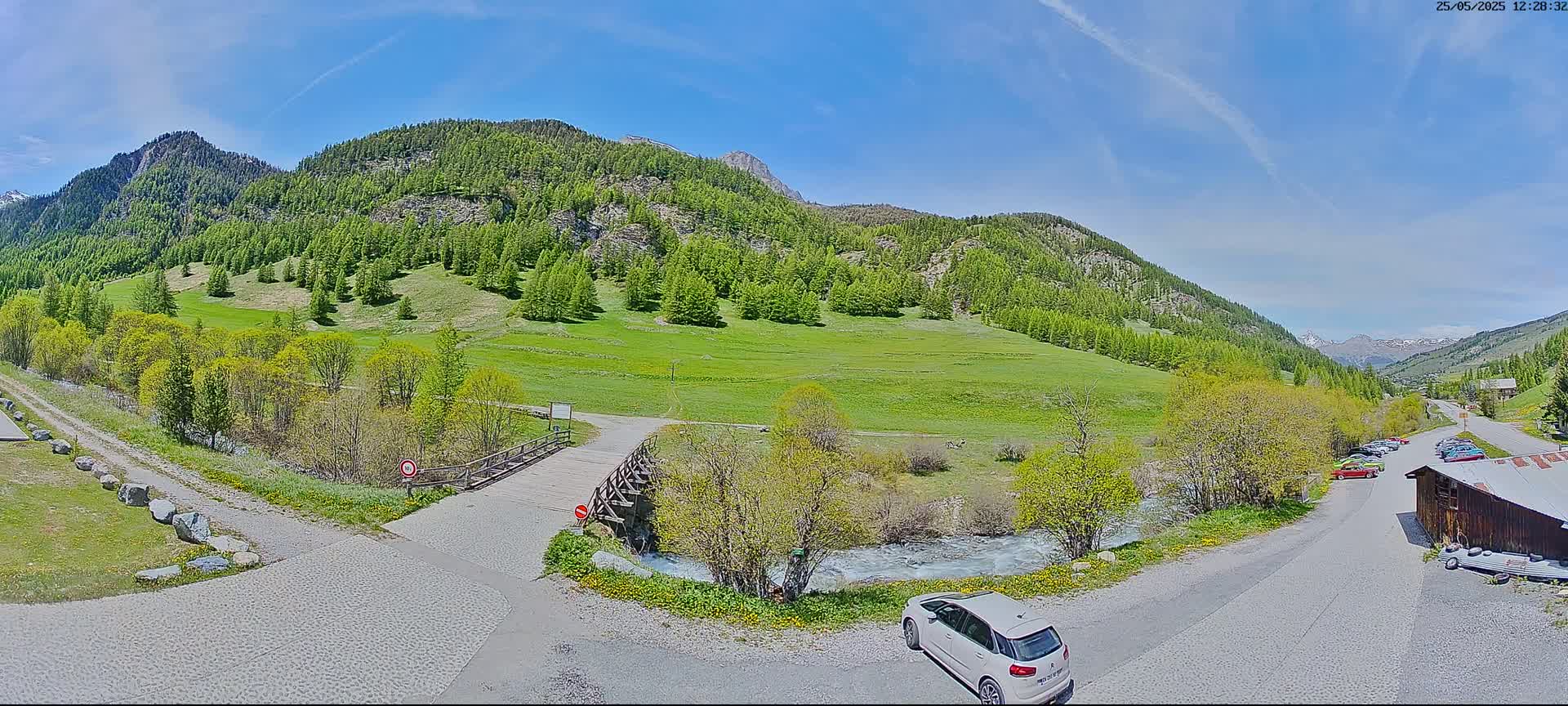 A white car is parked near a small wooden bridge over a stream, with a grassy valley and mountains in the background under a mostly sunny sky.