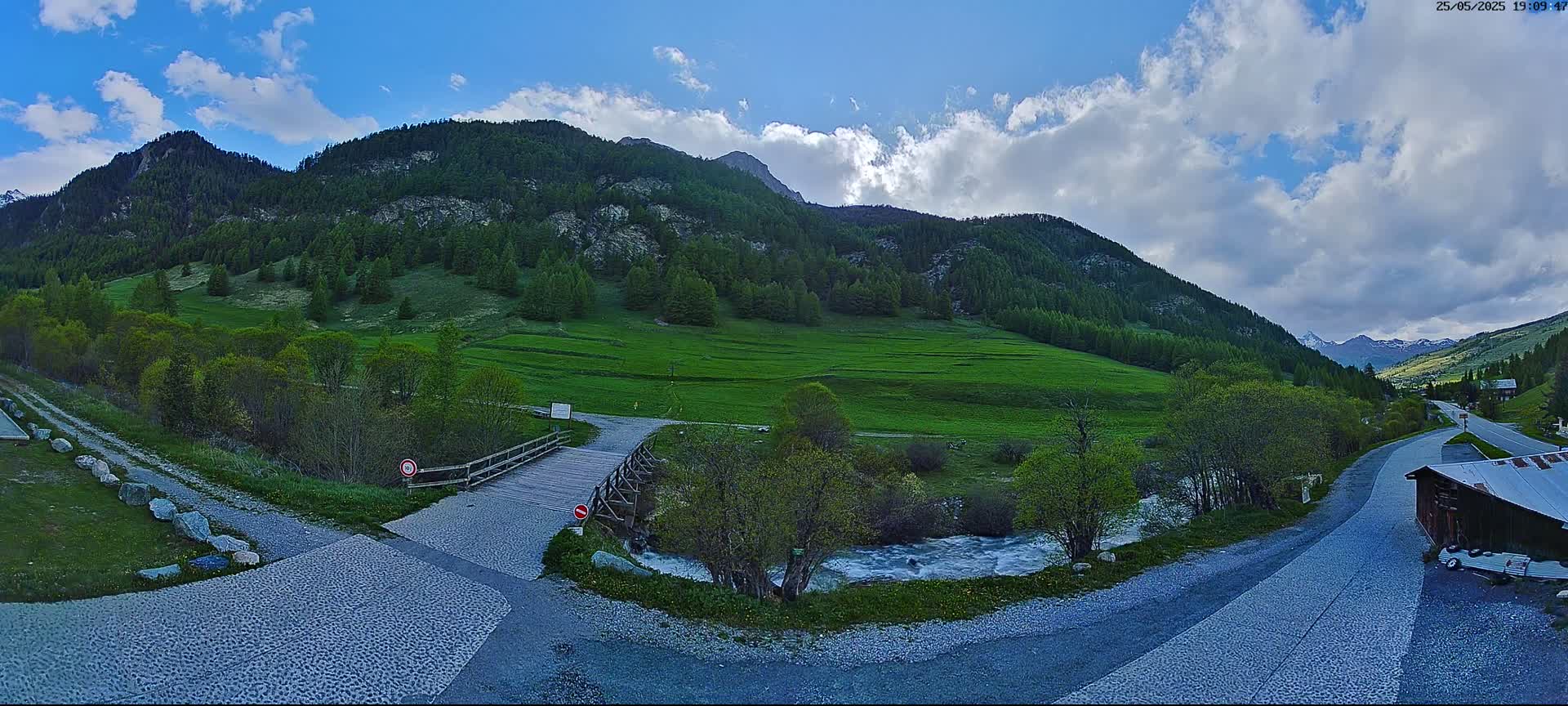 A wooden bridge crosses a small stream in a green valley nestled between mountains under a partly cloudy sky.
