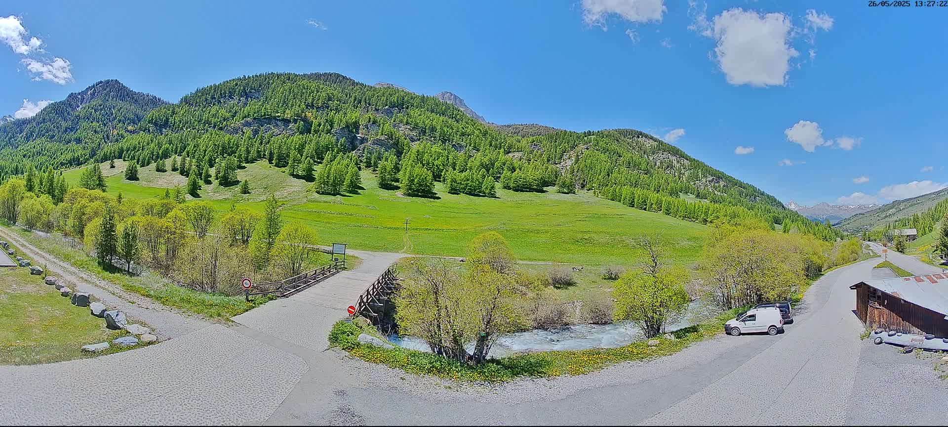 A sunny day in a mountainous valley shows a small wooden bridge over a stream, with green fields and forests, and a road leading to a small building.