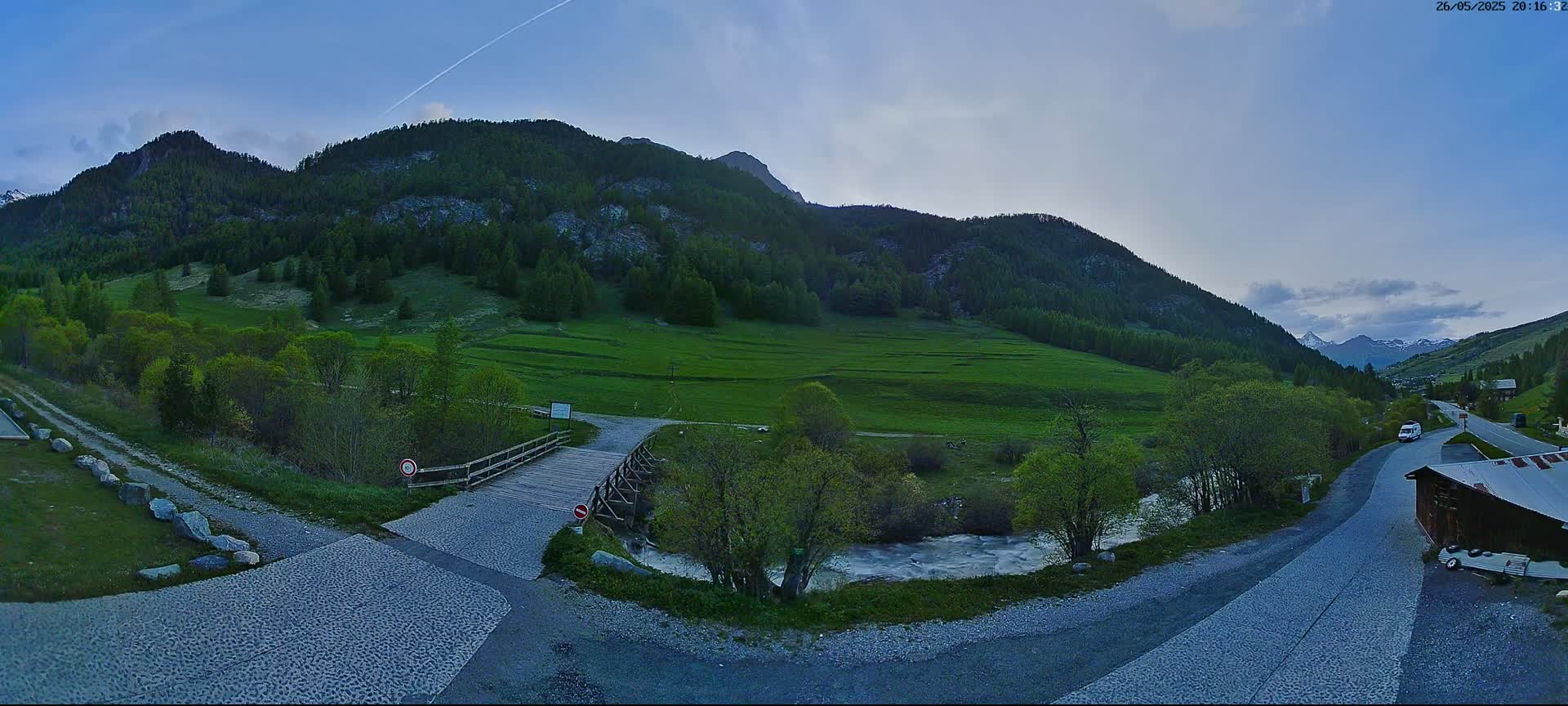 A wooden bridge crosses a river in a valley surrounded by green hills and mountains under a mostly clear, twilight sky.