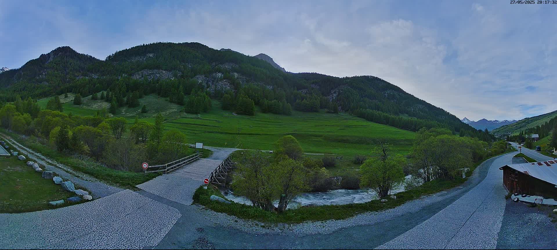 A wooden bridge crosses a small river in a green valley surrounded by mountains under a partly cloudy sky.