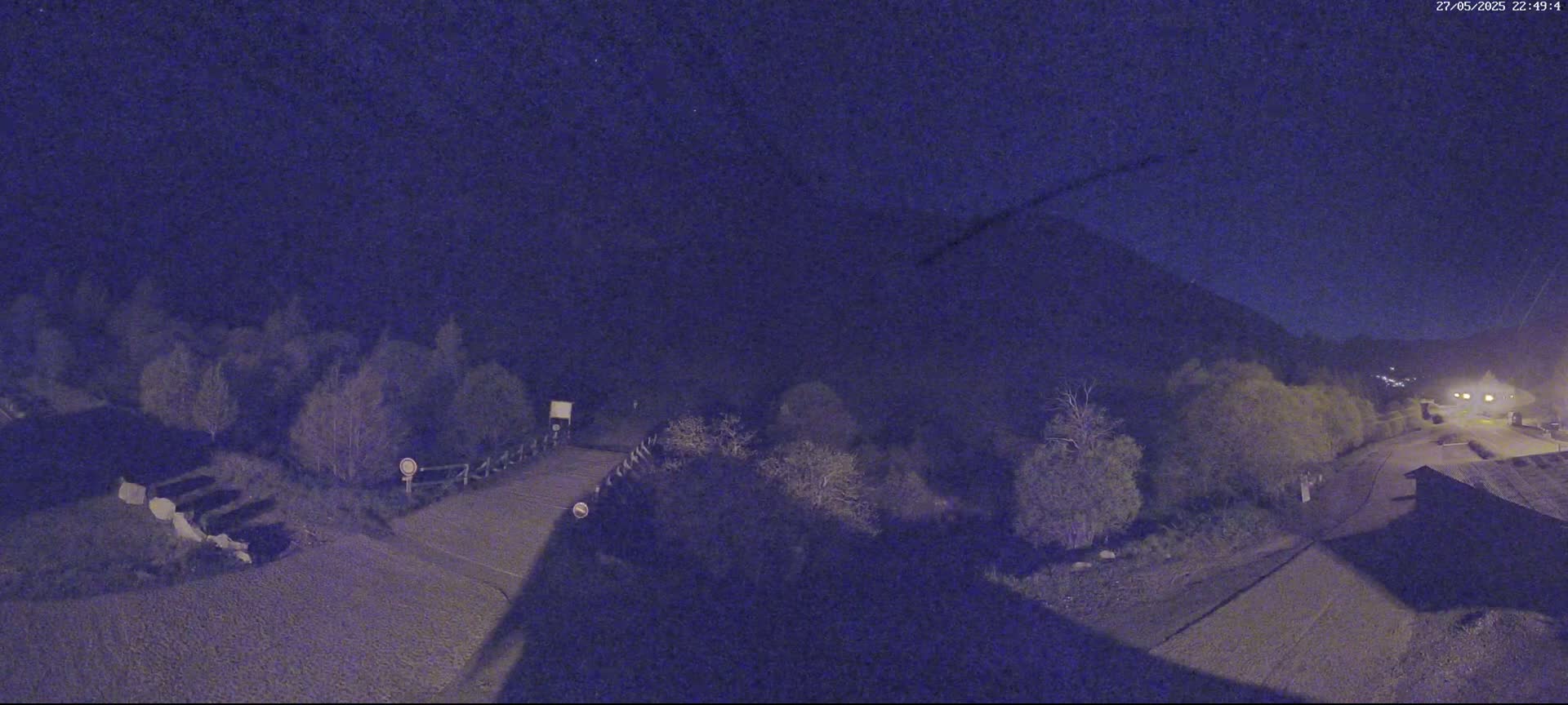A nighttime, high-angle view shows a road curving through a sparsely wooded area, under a dark, mostly clear sky.