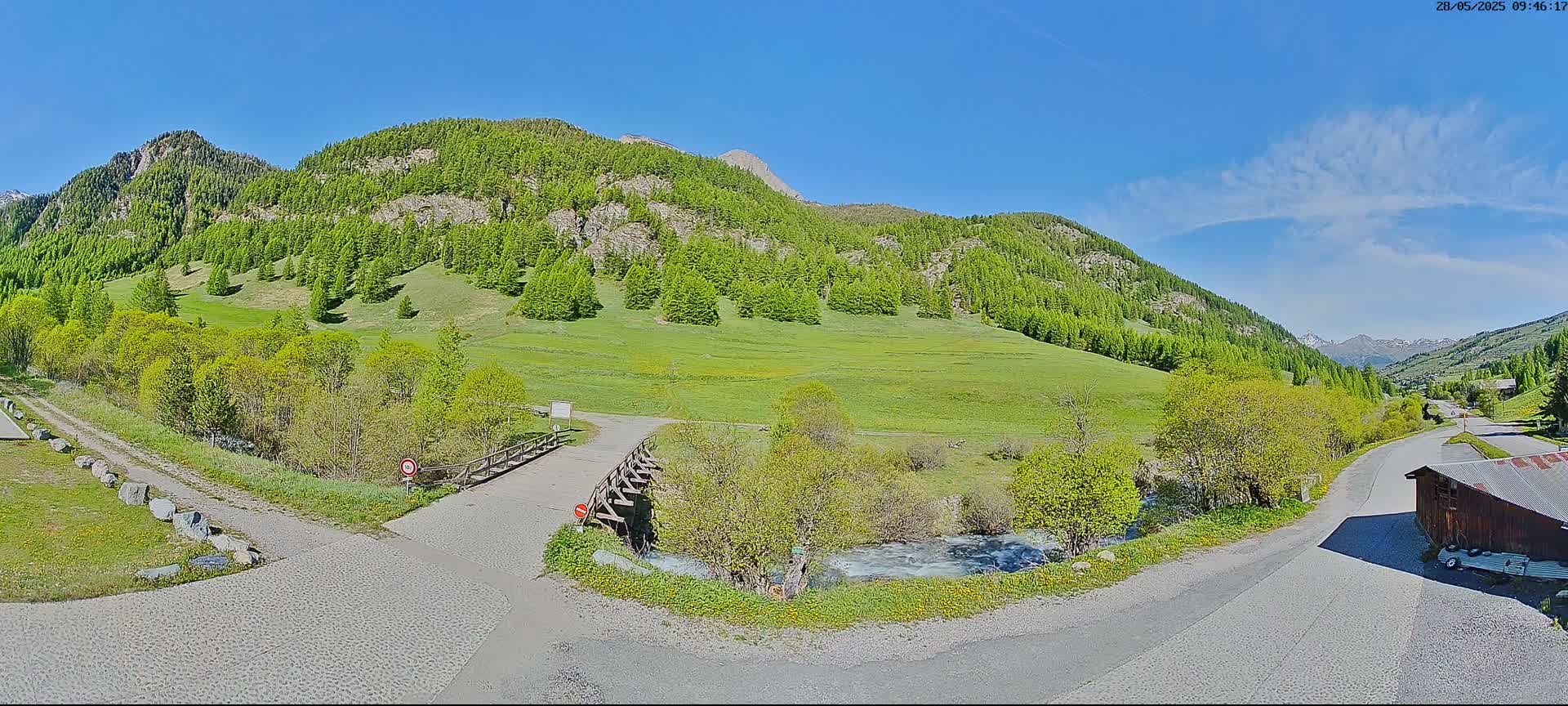 A sunny day in a mountainous region shows a valley of green grass and trees, a small stream, a wooden bridge, and roads leading to a small building.