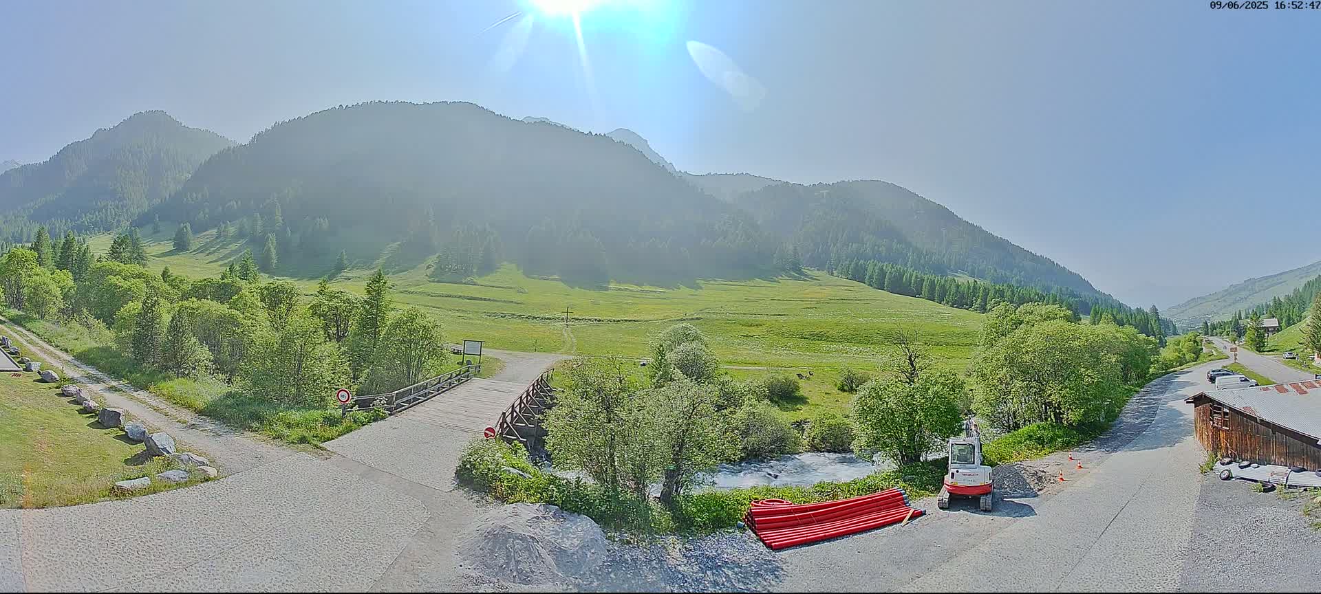 A sunny day in a mountainous valley reveals a small wooden bridge over a stream, a small construction vehicle, and a pile of red pipes near a gravel road and lush green vegetation, with mountains in the background.