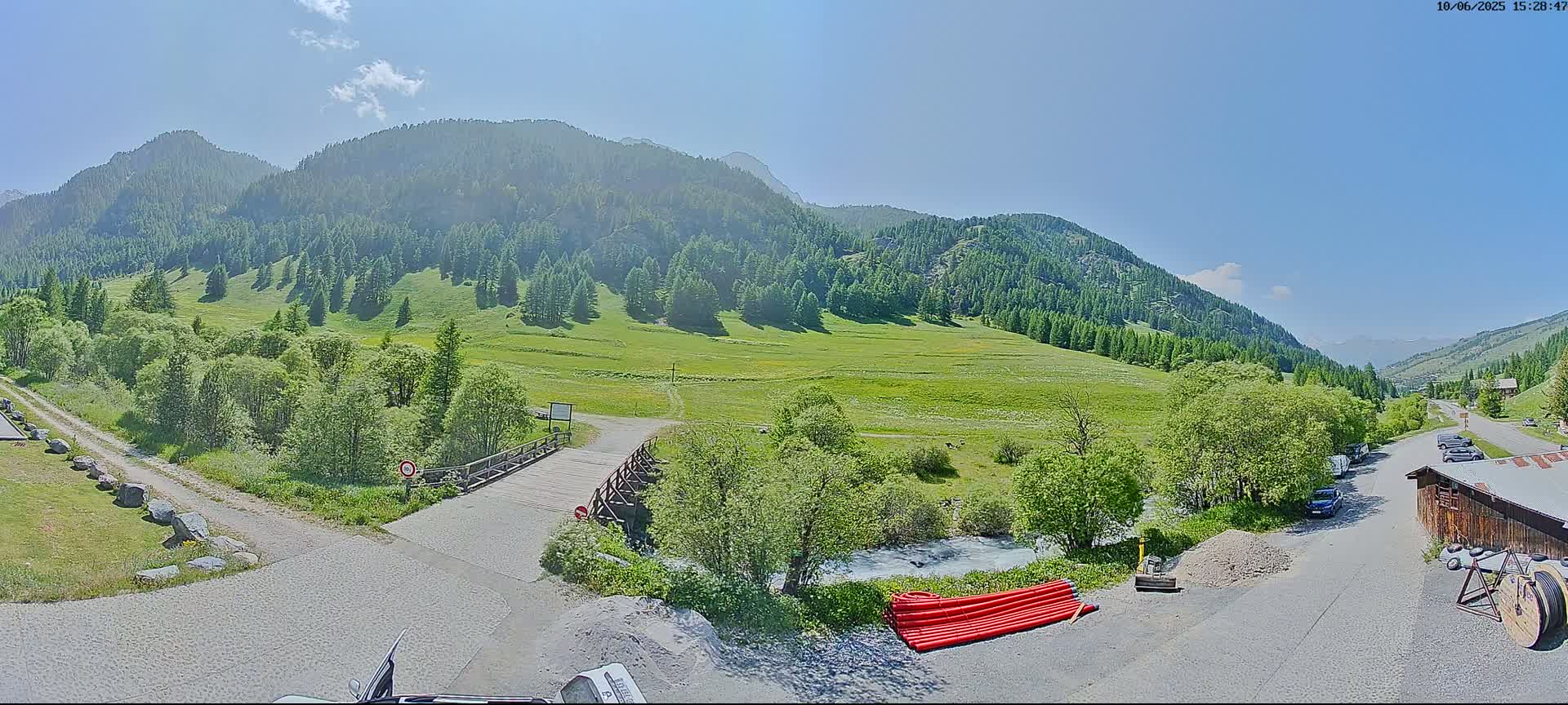 A sunny day reveals a mountainous landscape with green hills, a small wooden bridge over a stream, and a gravel road leading to a parking area near a building.