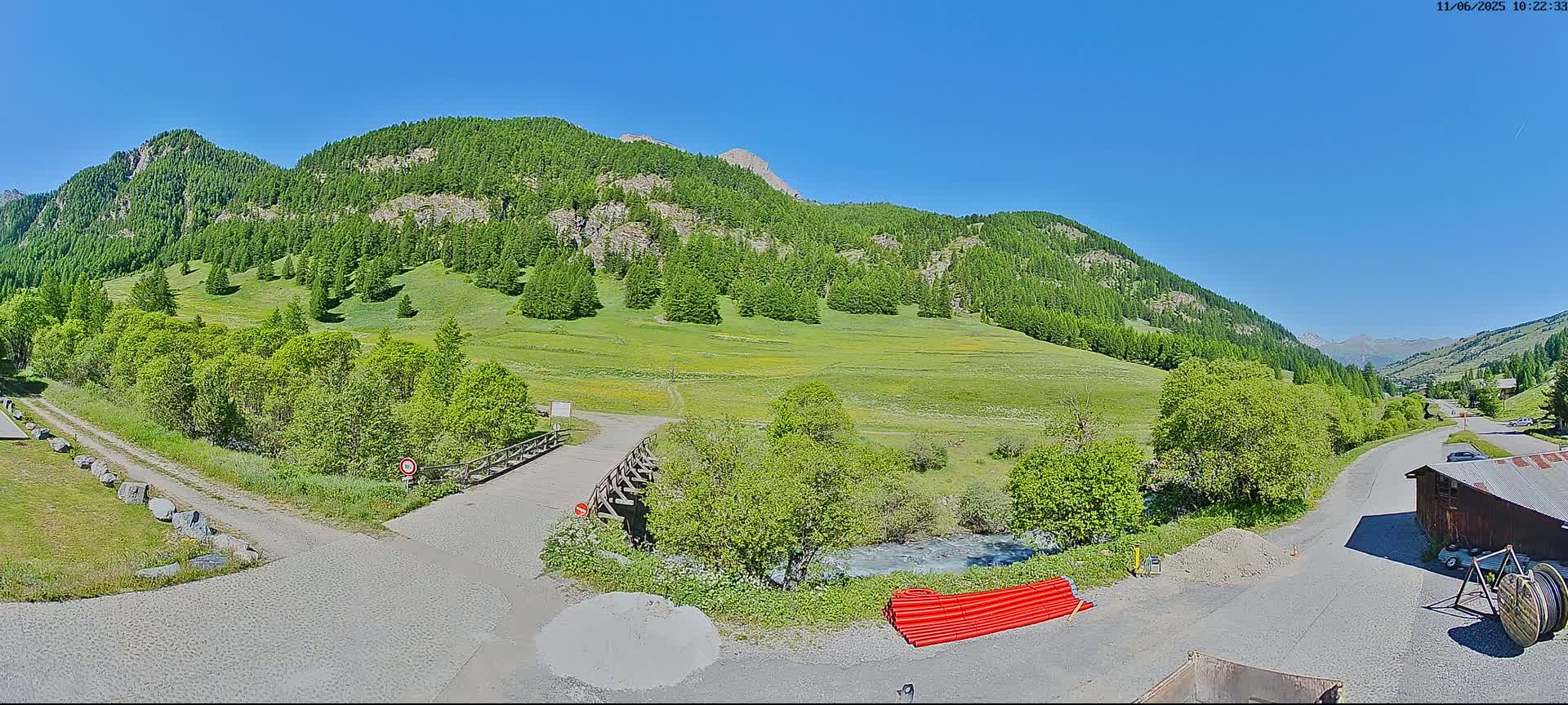 A sunny day reveals a valley scene with lush green meadows, coniferous-covered mountains, a small wooden bridge, and a road leading to a building.