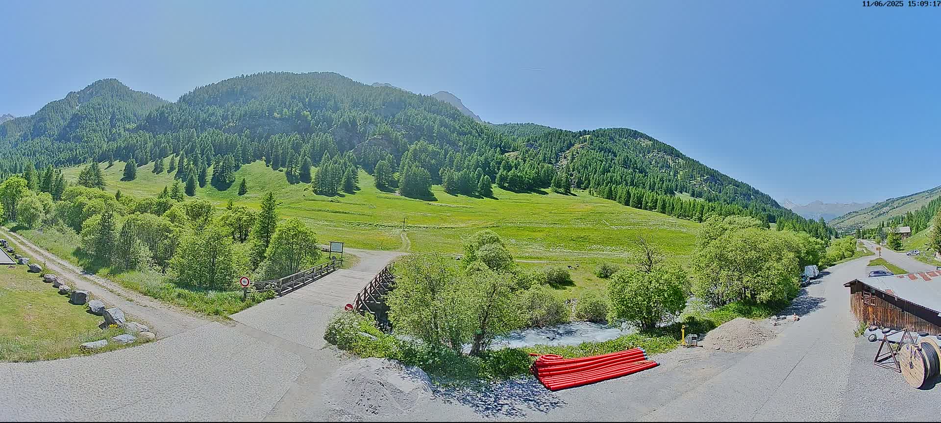A mountain valley under a clear blue sky features a small wooden bridge over a stream, with lush green meadows and coniferous forests on the hillsides.