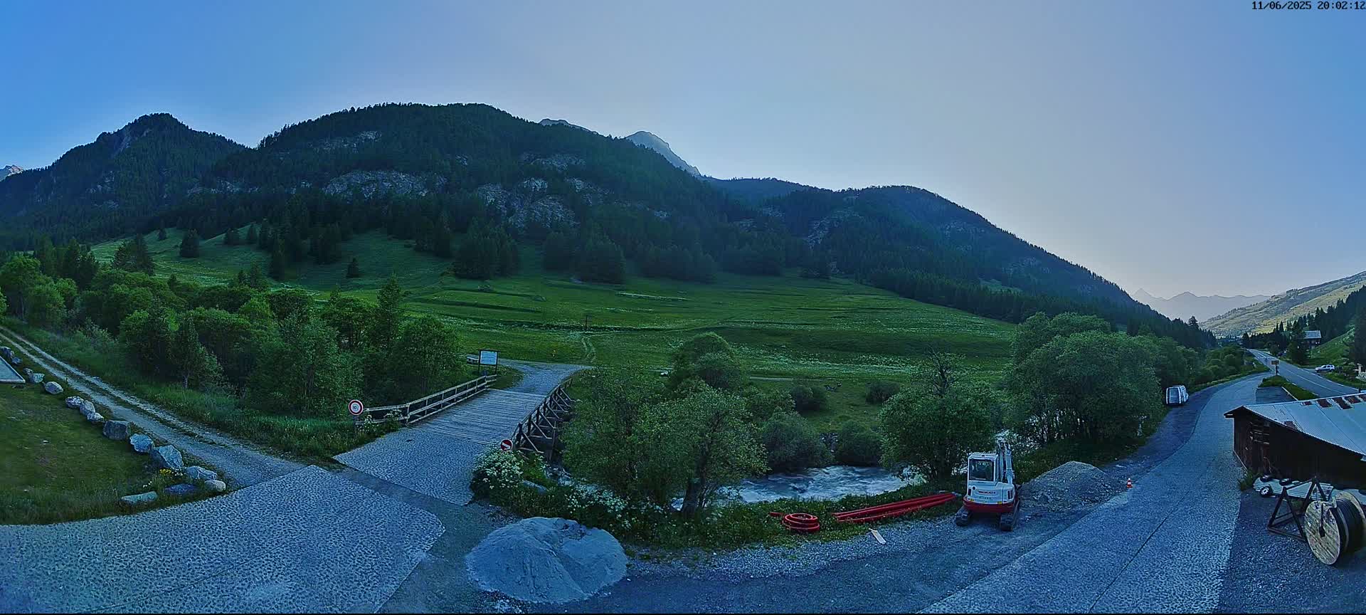 A small wooden bridge crosses a stream in a valley surrounded by lush green mountains under a clear blue sky.