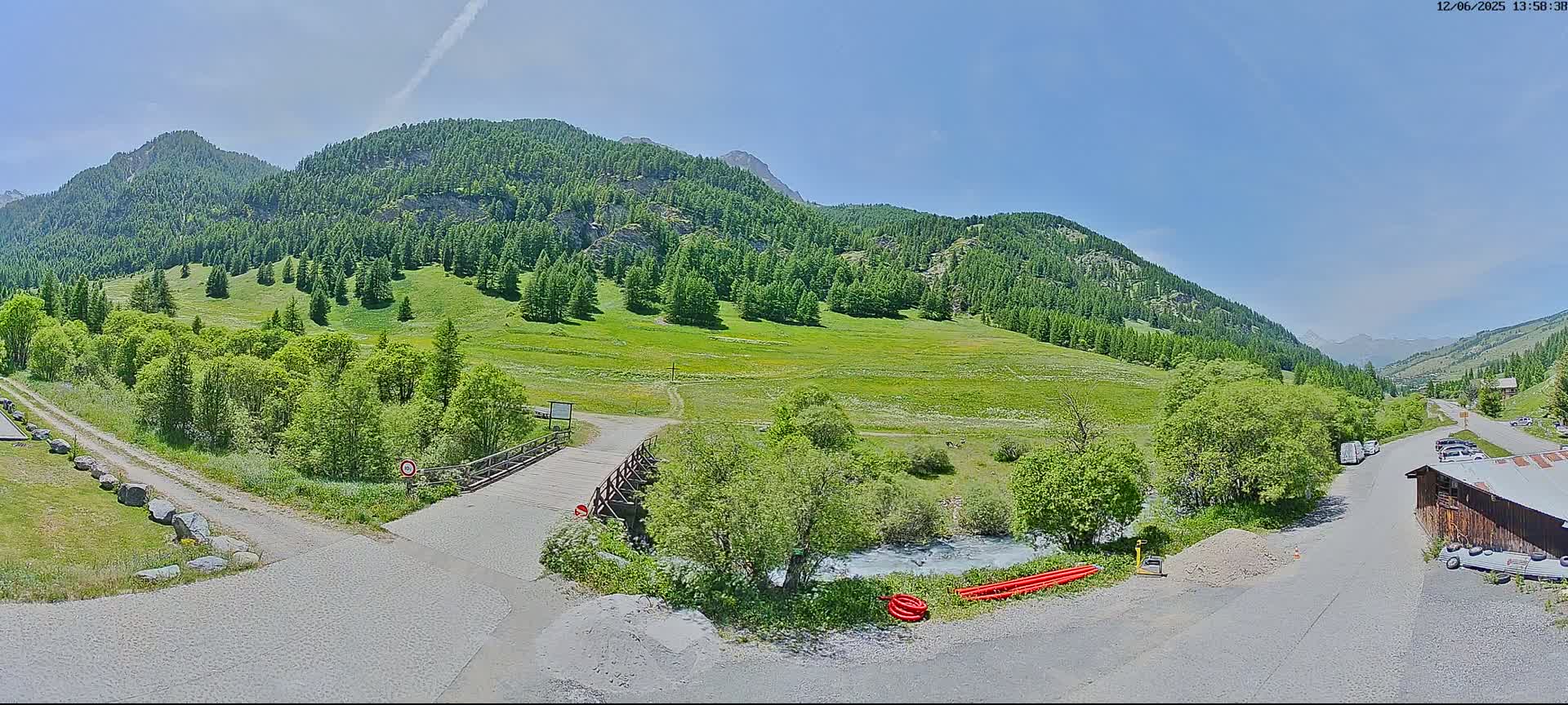 A wooden bridge crosses a small river in a lush green valley surrounded by mountains under a clear blue sky.