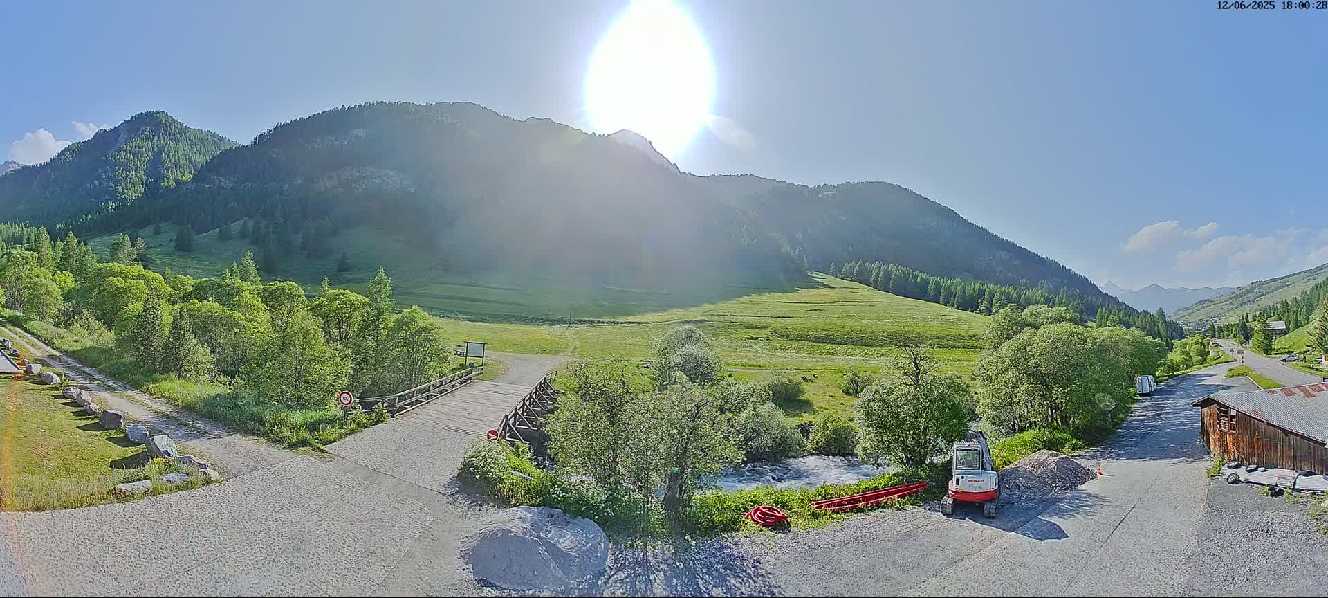 A sunny day reveals a mountainous landscape with a valley of green grass, trees, a small river, a wooden bridge, and a small construction vehicle parked near a building.