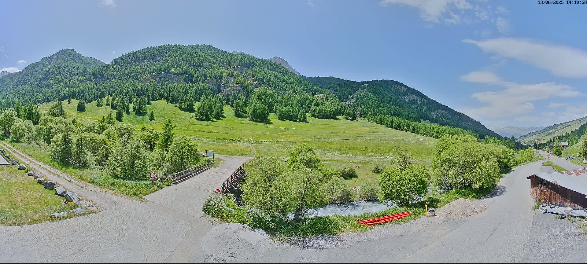 A sunny day reveals a valley scene with a small wooden bridge over a stream, green meadows, and coniferous trees covering the surrounding mountains.