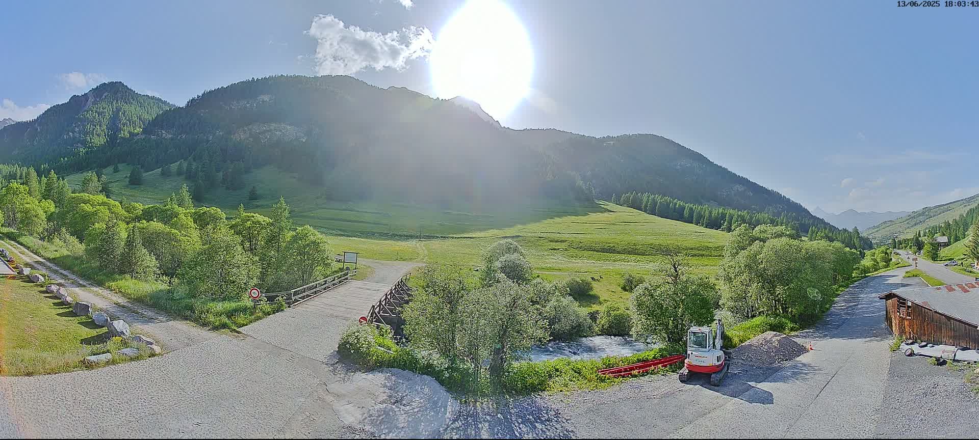A sunny day in a mountainous region shows a small bridge over a stream, a gravel road, and a small excavator parked near a building, with green fields and forests in the valley below the mountains.