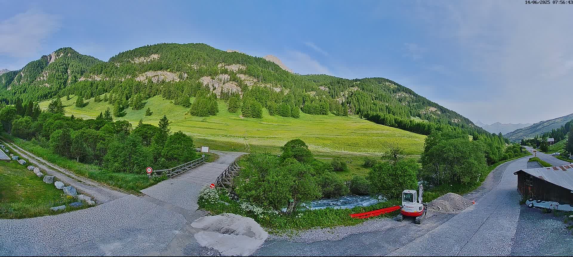 A sunny day in a mountainous region shows a small bridge over a stream, a gravel road, and a small excavator parked near a building, with green fields and forests in the valley below the mountains.