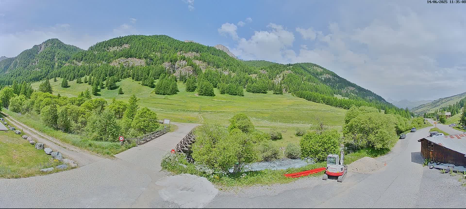 A sunny day in a mountainous valley shows a small wooden bridge over a stream, leading to a dirt road that winds past a grassy field and up toward a lush, green mountainside.