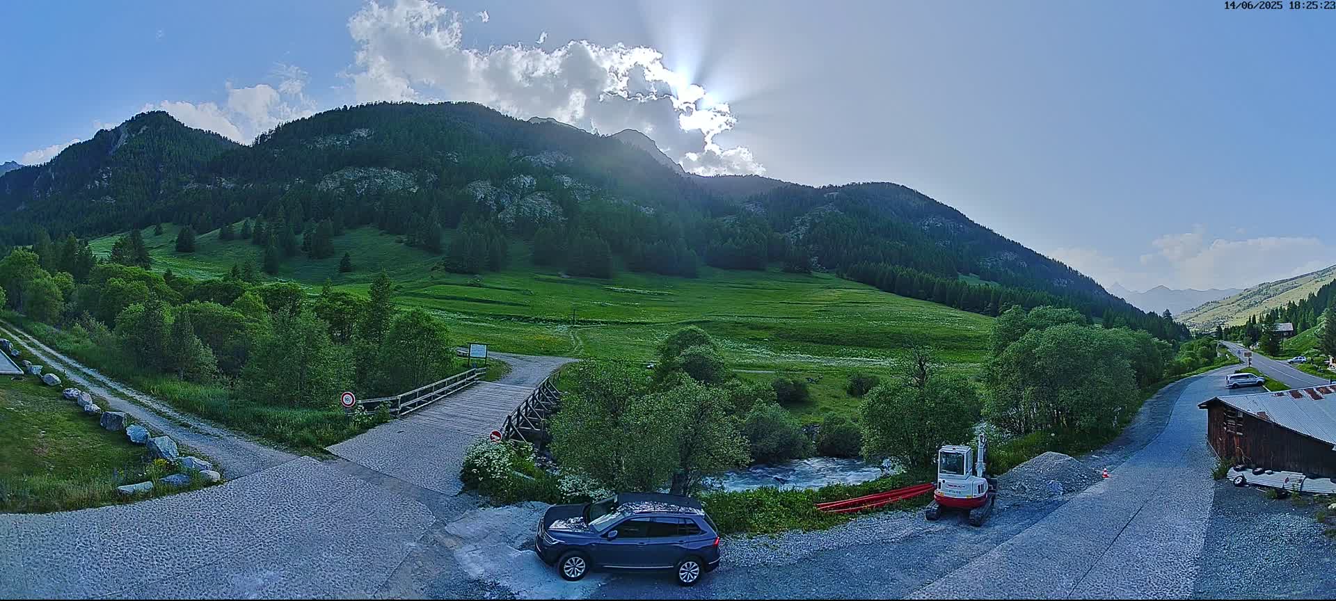 A sunny day in a mountainous valley shows a small wooden bridge over a stream, a dark blue SUV parked nearby, a small excavator, and a gravel road leading to a building.
