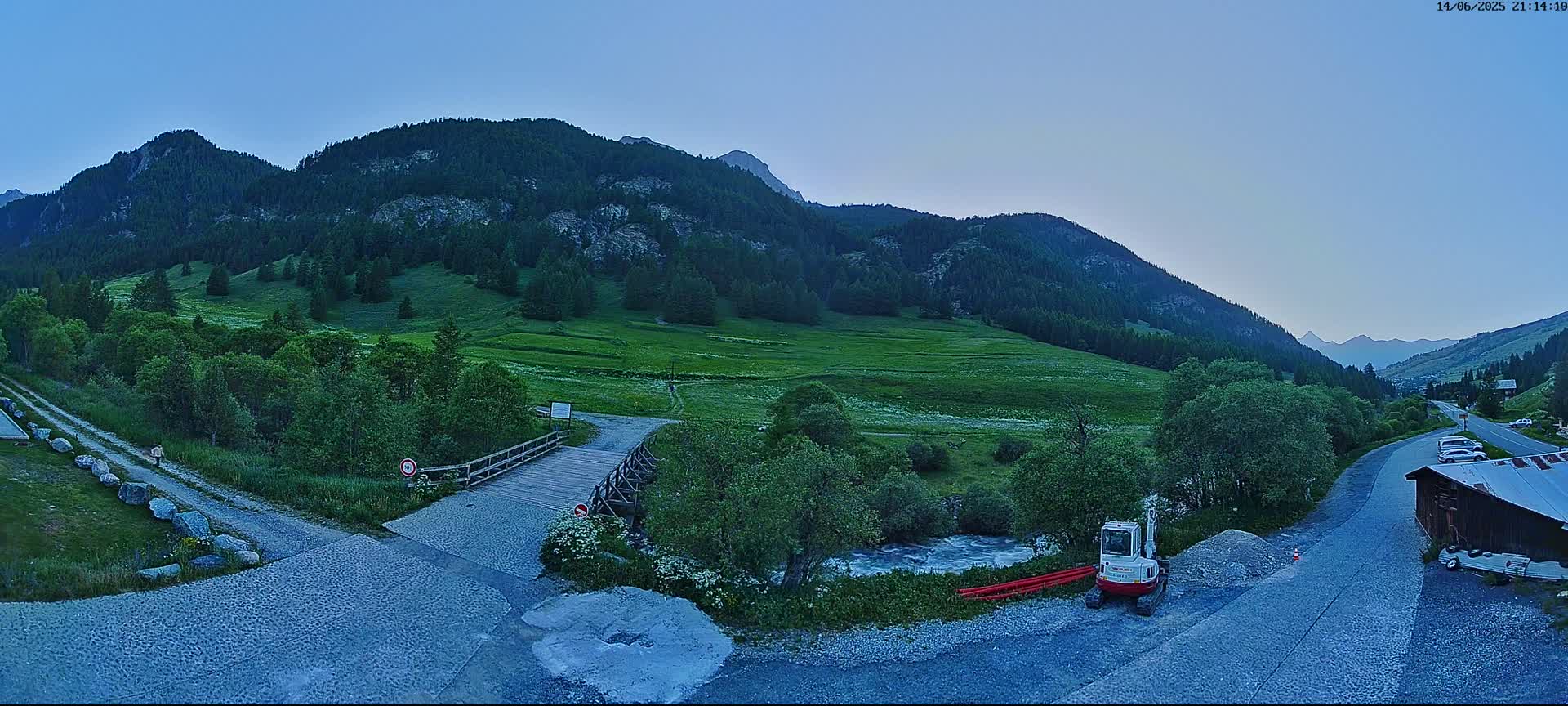 A sunny day in a mountainous valley shows a small wooden bridge over a stream, a dark blue SUV parked nearby, a small excavator, and a gravel road leading to a building.