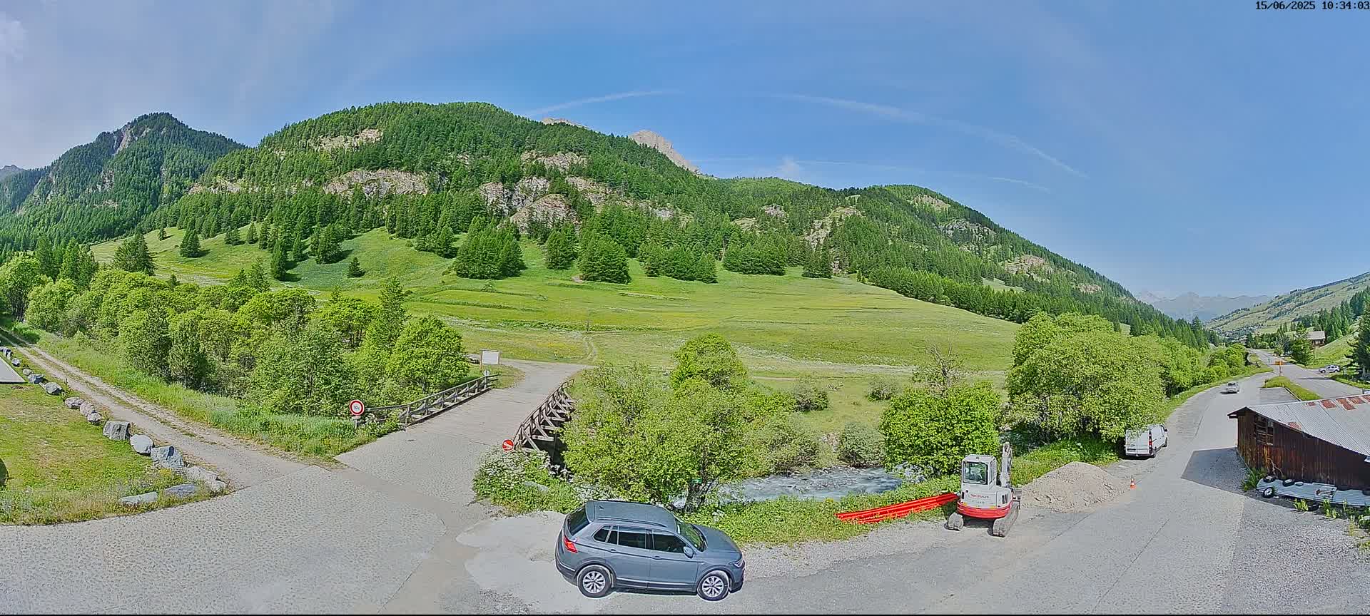 A gray SUV is parked near a small bridge over a stream in a mountainous valley under a clear, sunny sky.