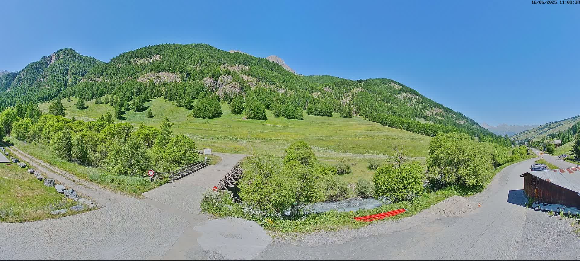 A mountain valley on a sunny day shows a small wooden bridge over a stream, roads, and lush green vegetation.