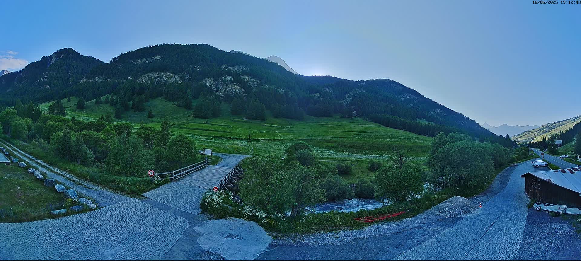 A wooden bridge crosses a small stream in a valley surrounded by lush green mountains under a clear blue sky.