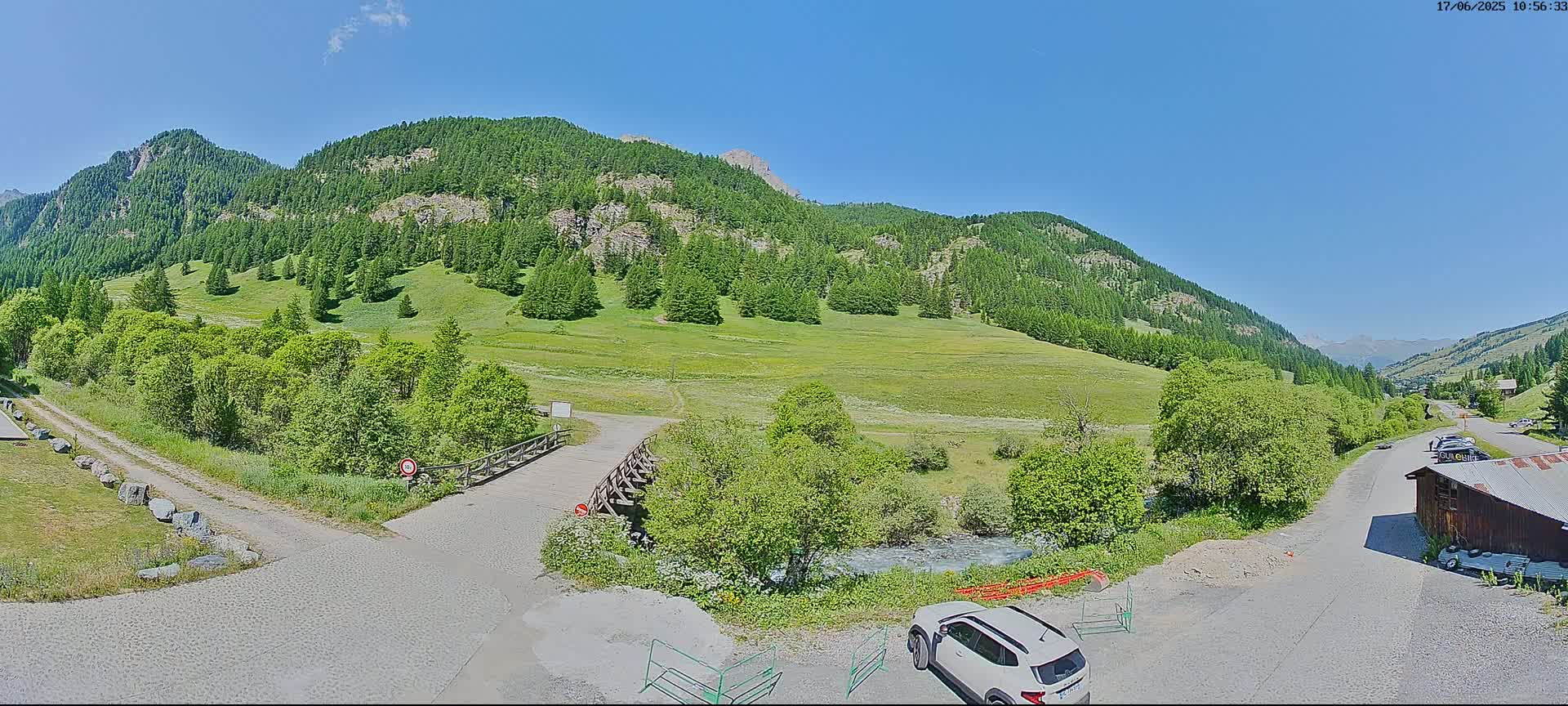 A mountain valley scene on a sunny day shows a paved road leading to a wooden bridge over a small stream, with lush green vegetation, a white SUV parked nearby, and distant mountains under a clear blue sky.
