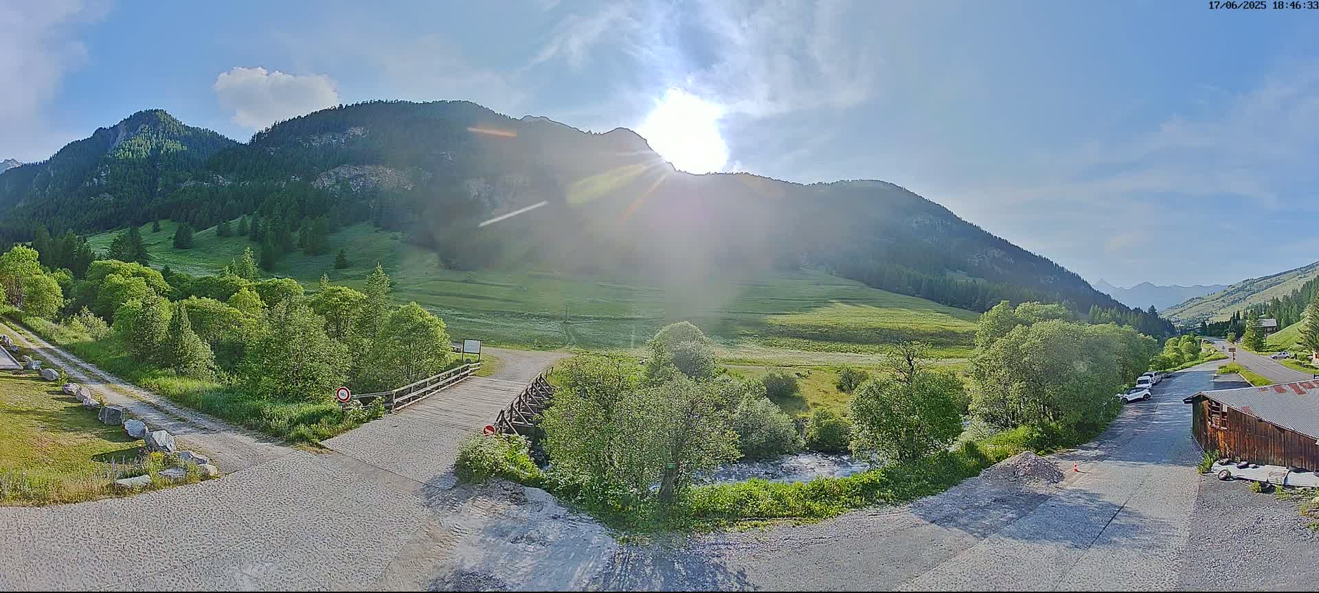 A sunny day in a mountainous valley shows a gravel road leading to a small bridge over a stream, with green meadows and forested hillsides surrounding a parking area with a few cars.