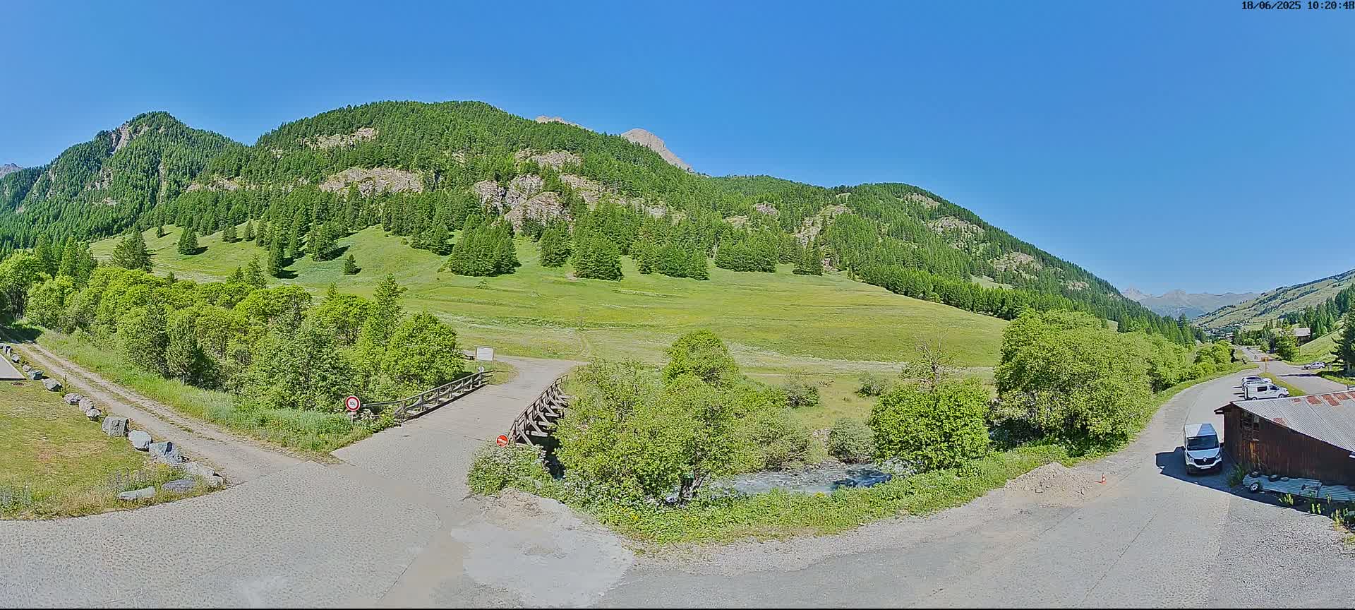 A mountain valley under a clear blue sky shows a dirt road leading to a small wooden bridge over a stream, with green meadows and evergreen forests covering the hillsides.