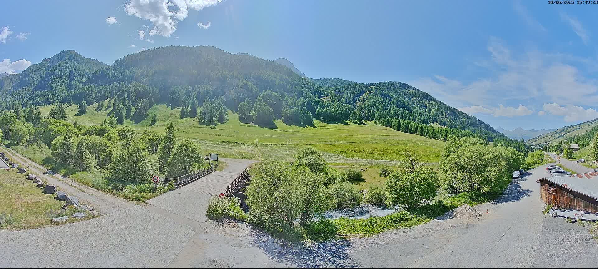 A sunny mountain valley scene features a small wooden bridge over a stream, gravel roads, green meadows, and coniferous forests on the hillsides.