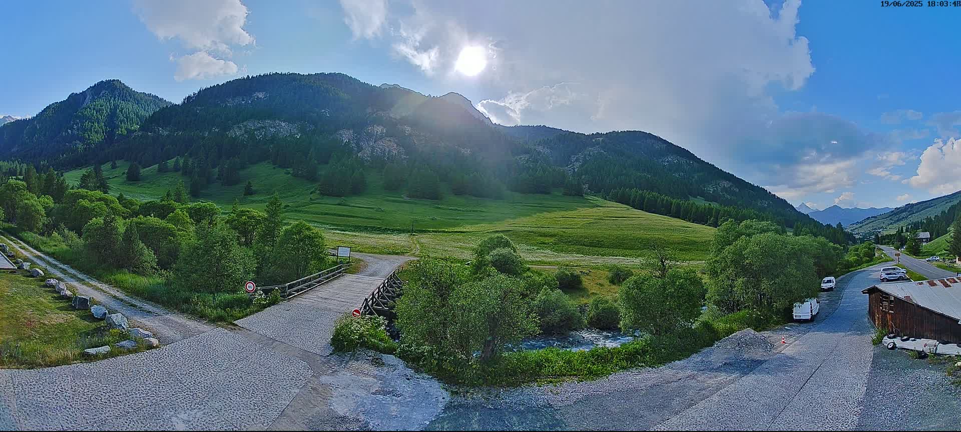 A wooden bridge crosses a small stream in a valley surrounded by lush green hills and mountains under a partly sunny sky.
