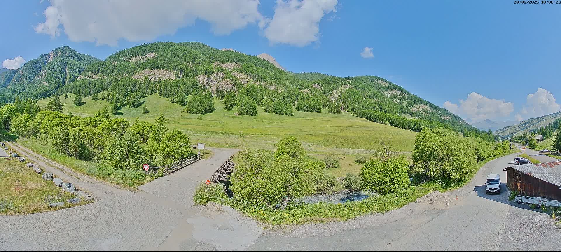 A partly sunny day reveals a mountainous landscape with green meadows, a small stream, a wooden bridge, and a dirt road leading to a paved road where a few vehicles are parked near a rustic building.