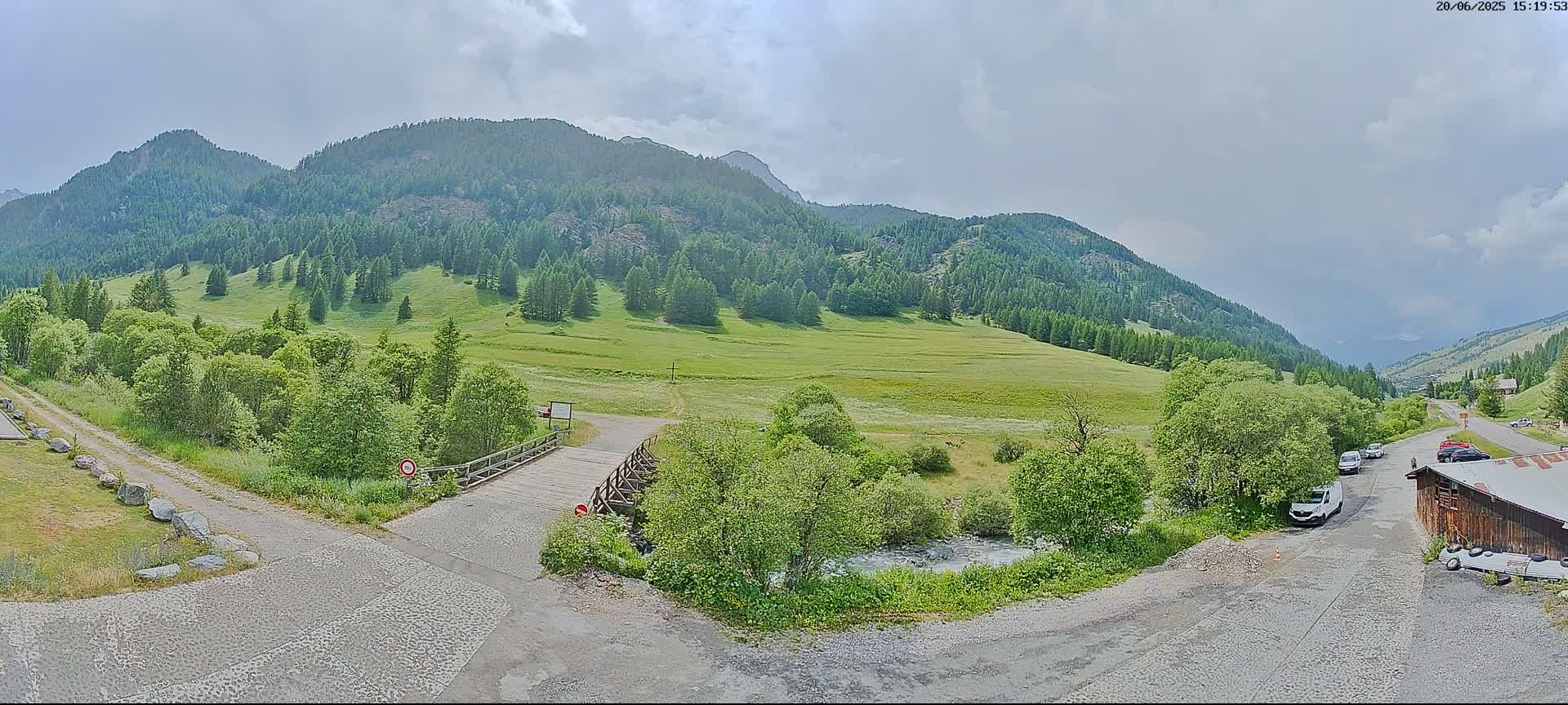 A wooden bridge crosses a small stream in a valley surrounded by green, forested mountains under a partly cloudy sky.
