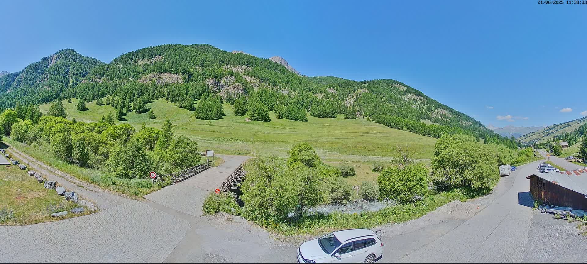 A sunny, mountainous landscape features a small wooden bridge over a stream, a paved road, and a white station wagon parked near a building.