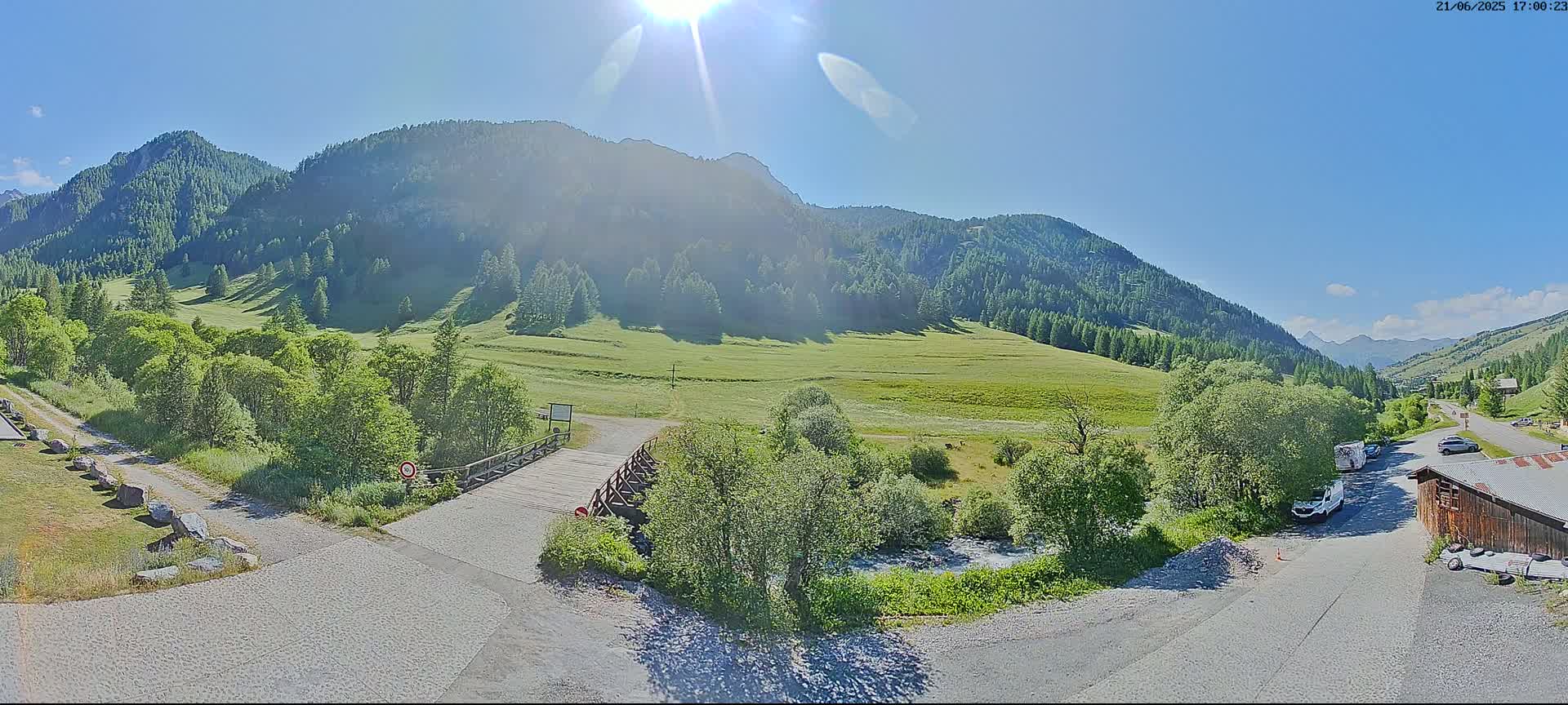 A sunny day in a mountainous valley shows a small bridge over a stream, with green meadows and trees, a gravel road, and a small parking area with a few vehicles.