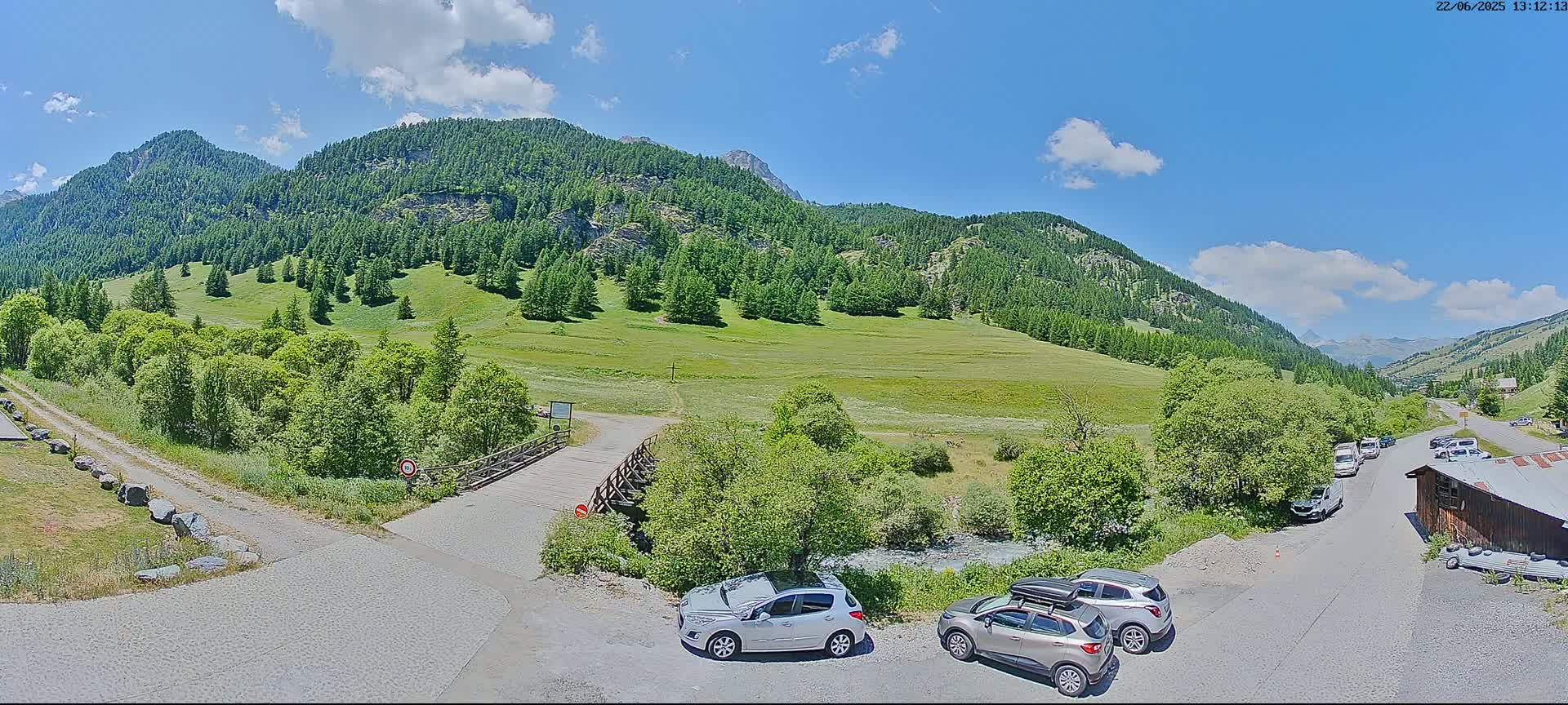 A sunny day reveals a mountainous landscape with green meadows, coniferous forests, a small wooden bridge, and several cars parked near a building.