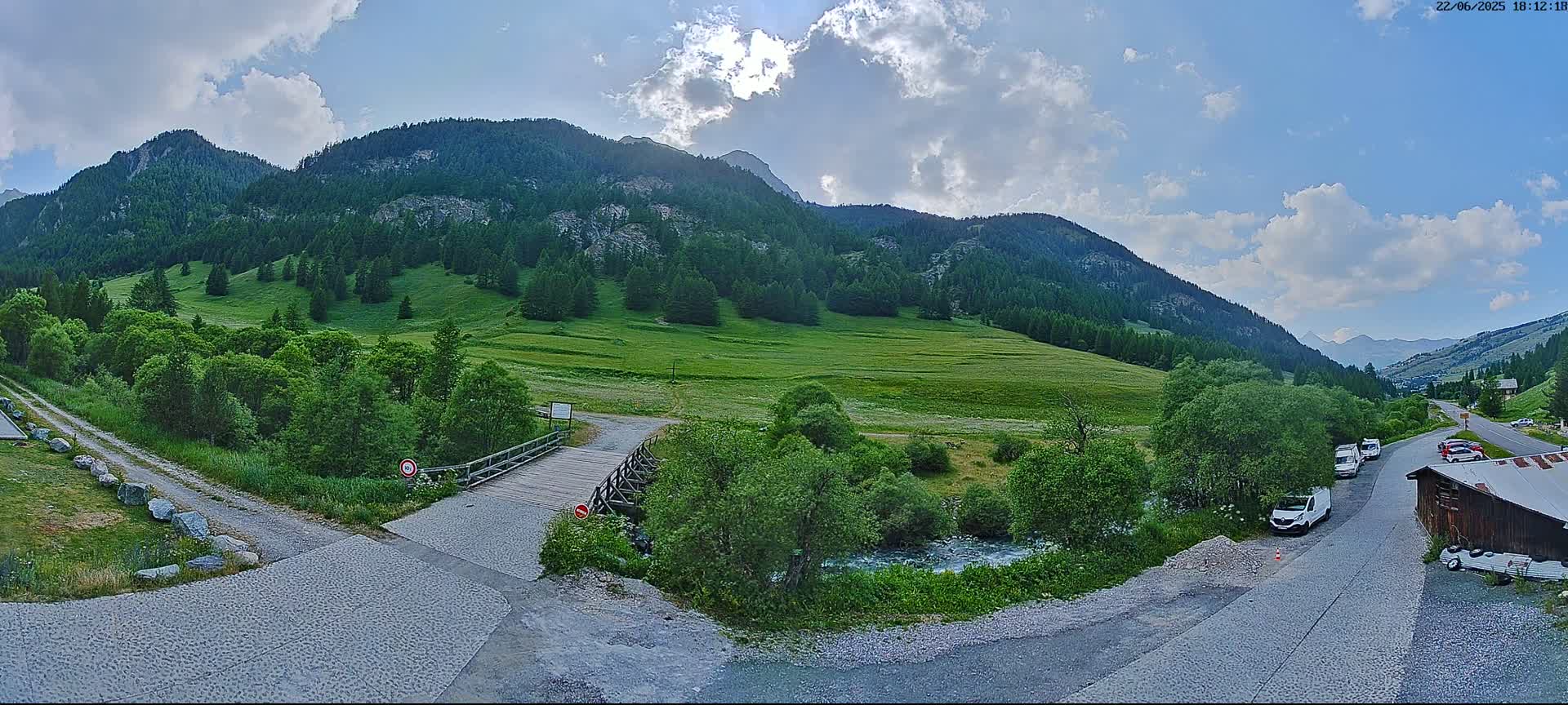 A partly cloudy day reveals a mountain valley with green fields, a small wooden bridge over a stream, and a gravel road leading to a rustic building; several vehicles are parked nearby.