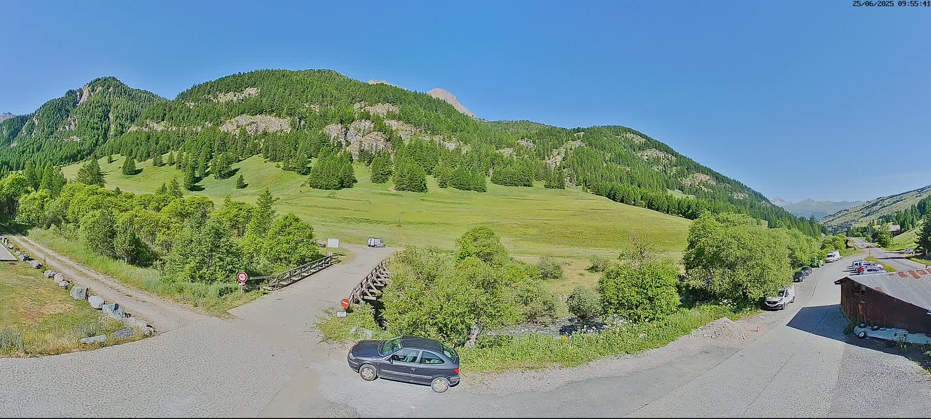 A sunny mountain valley scene with green meadows, evergreen trees, a small stream, parked cars, and a few buildings, under a clear blue sky.