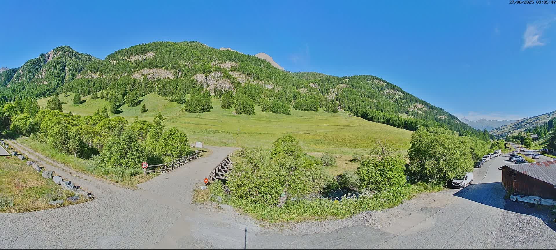 A sunny day in a mountainous region shows a grassy valley surrounded by lush green trees and rocky slopes, with a paved road leading to a parking area with several cars.