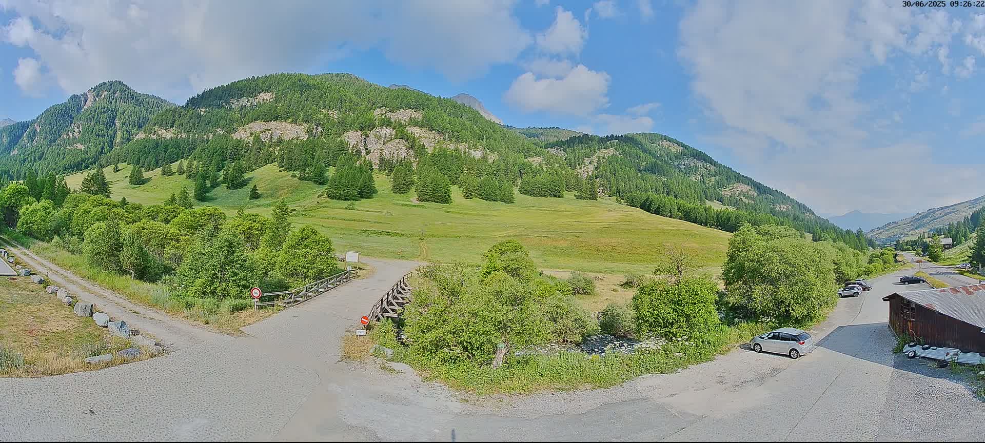 A mountain valley with a grassy field and a few cars parked near a small building under a partly cloudy sky.