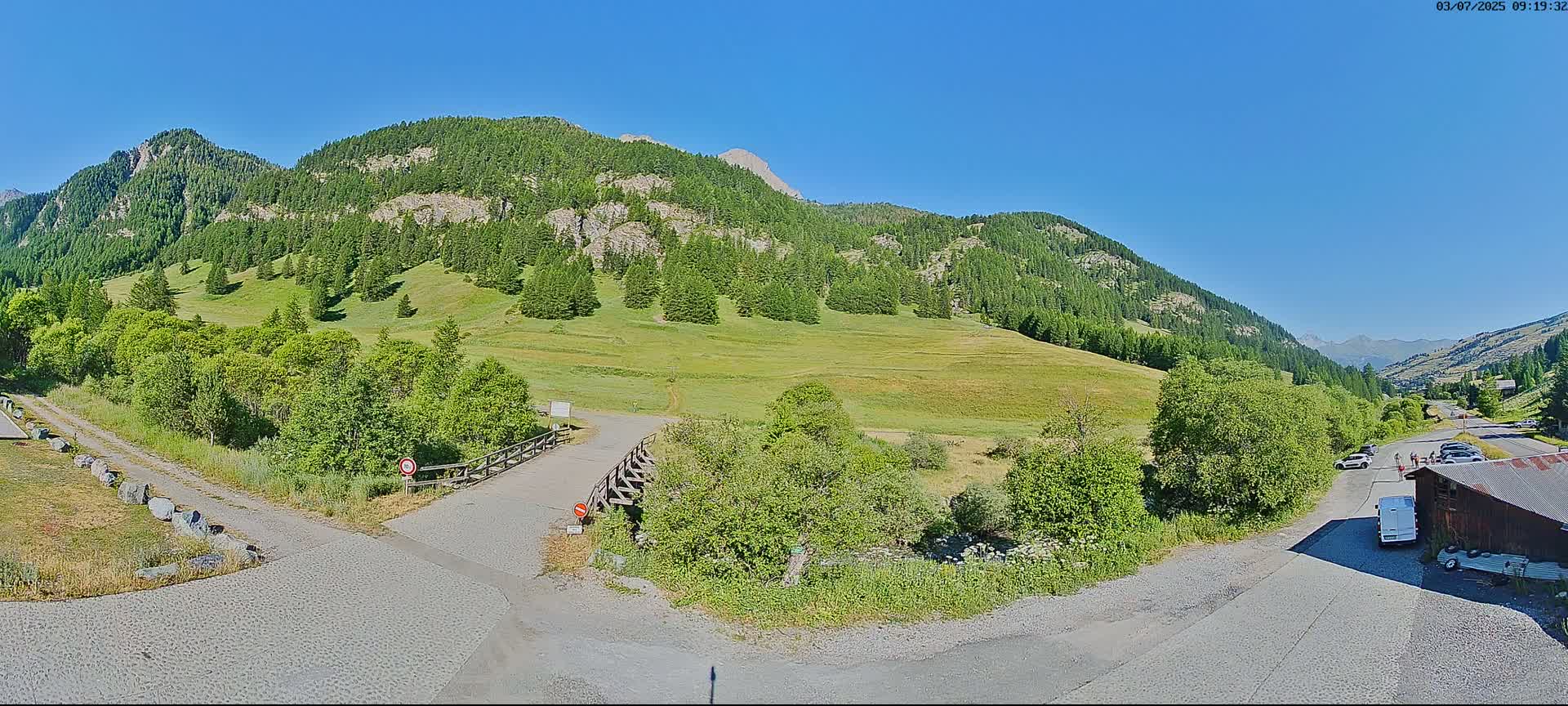 A paved road leads to a small wooden bridge crossing a grassy area, with lush green mountains and a clear blue sky in the background.