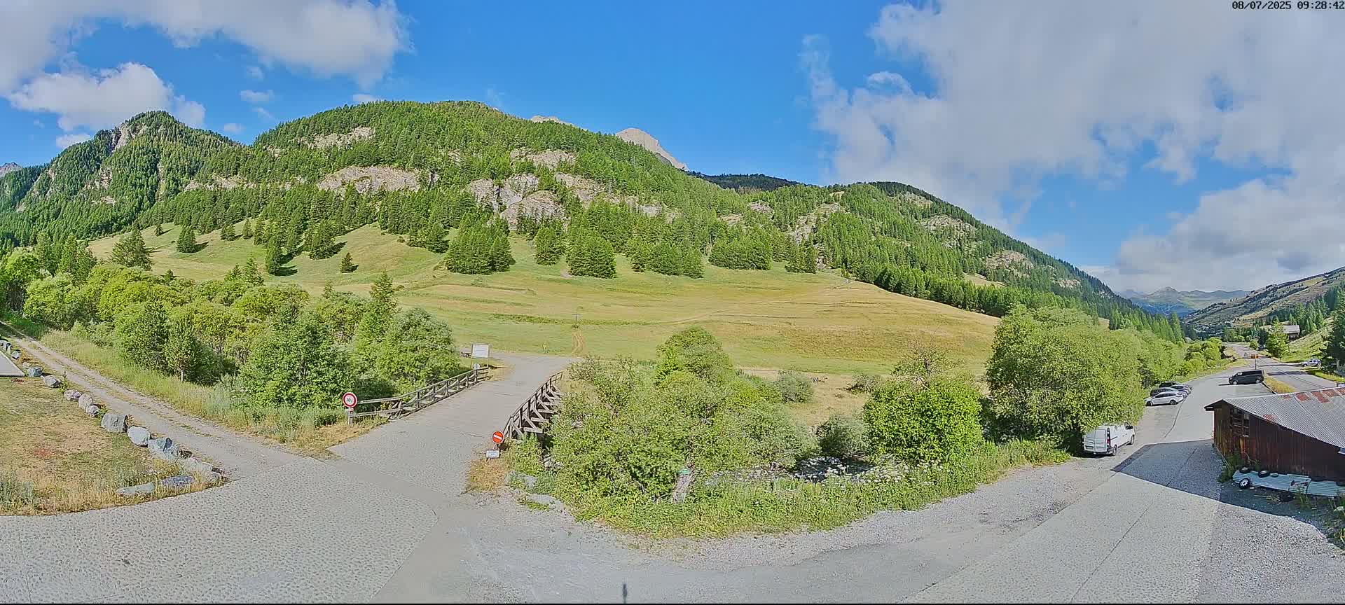 A partly cloudy sunny day reveals a mountain valley with a grassy field, green trees, a small parking area with several vehicles, and a gravel road leading to a wooden structure.