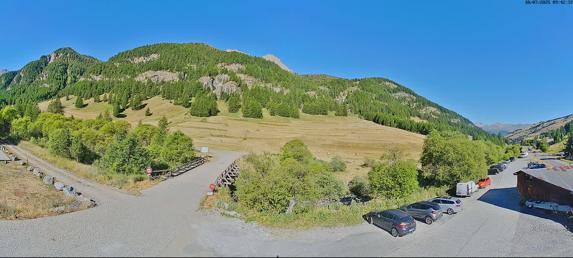 A sunny mountain valley scene with a gravel road, several parked cars, and a wooden building, all set against a backdrop of green hills and a clear blue sky.