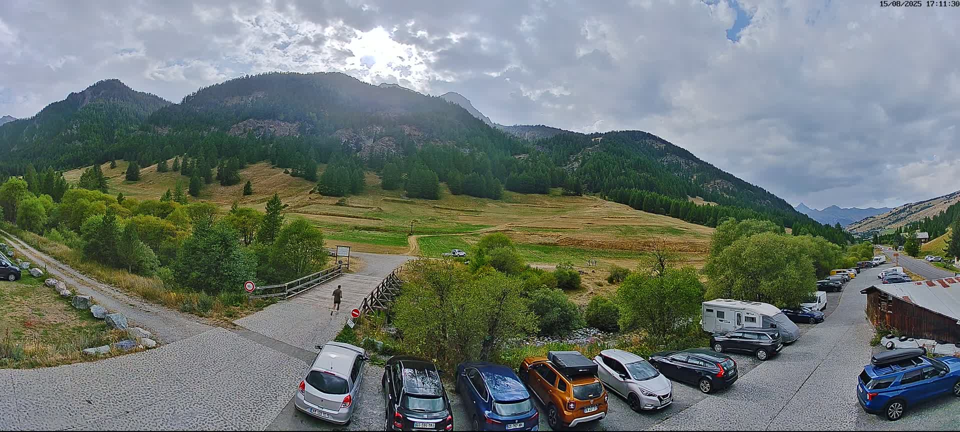 A sunny mountain valley scene with a gravel road, several parked cars, and a wooden building, all set against a backdrop of green hills and a clear blue sky.