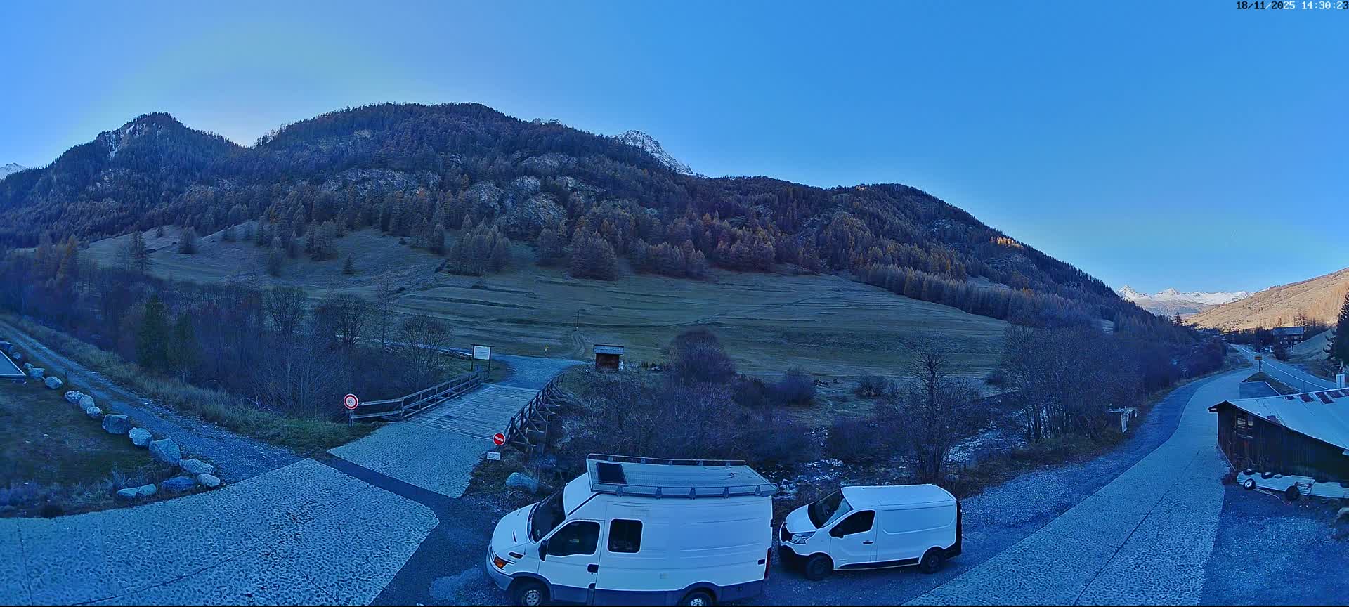 A sunny mountain valley scene with a gravel road, several parked cars, and a wooden building, all set against a backdrop of green hills and a clear blue sky.